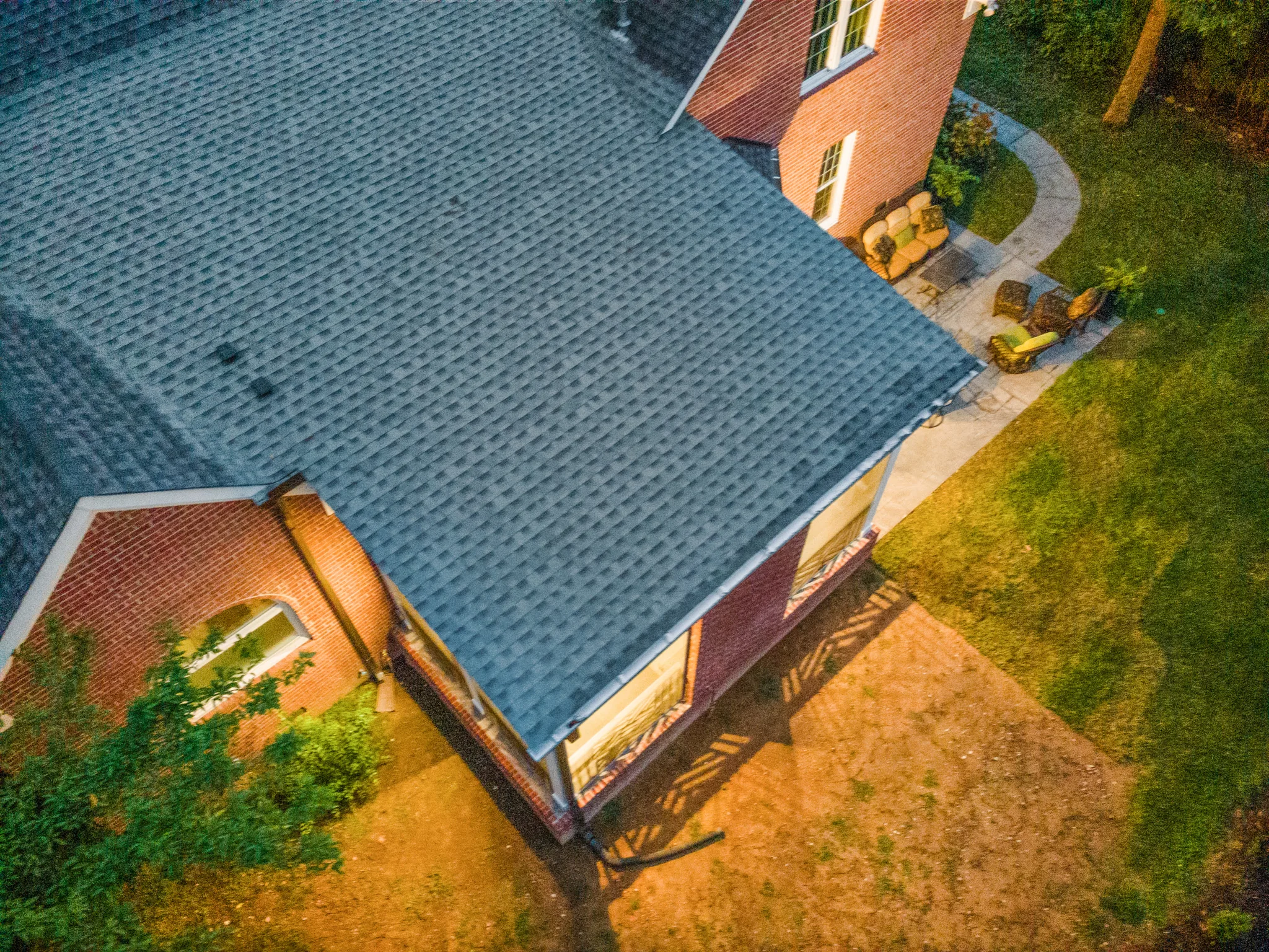 Aerial view of a brick home with gray asphalt shingle roof, curved concrete walkway, patio seating and a bare dirt yard