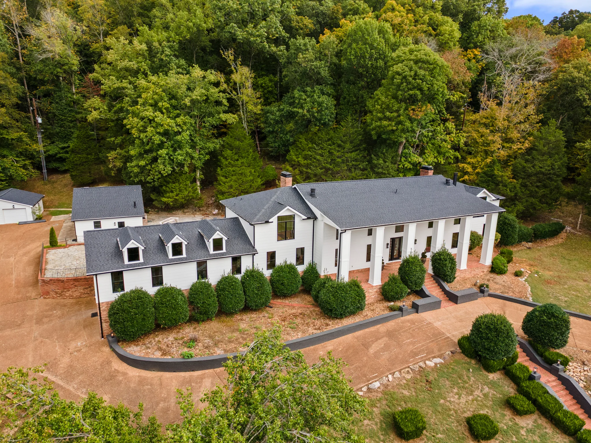 Aerial view of a long white home with black shingle roof, columned porch, dormered wing and clipped shrubs against a wooded hillside