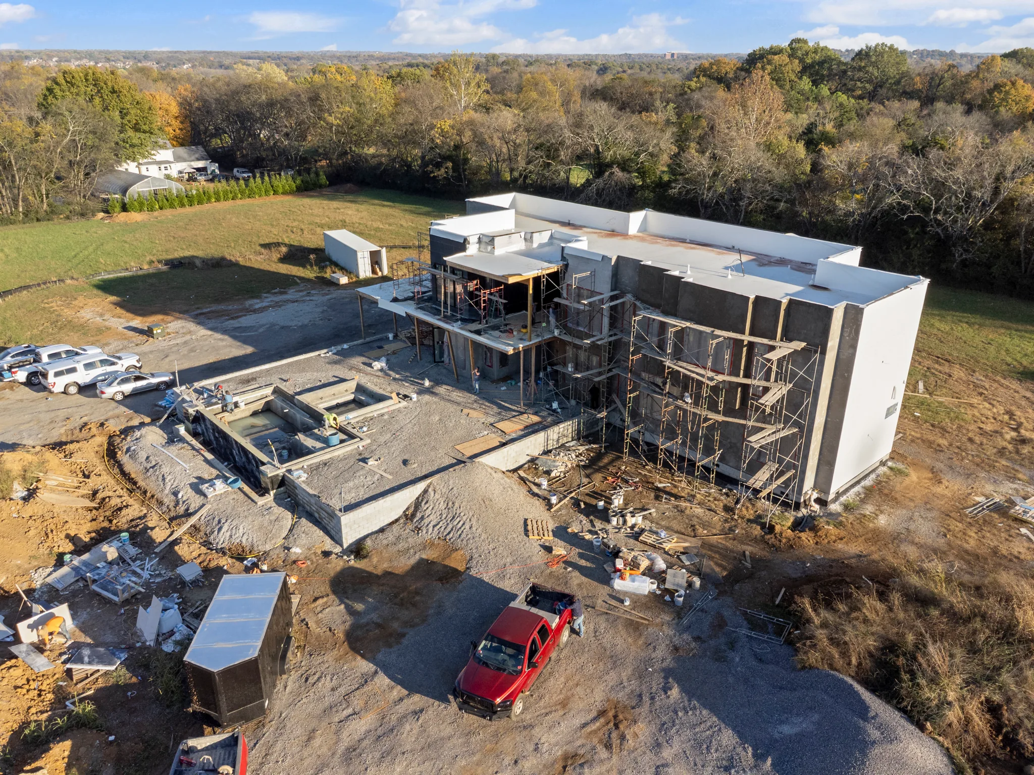Aerial view of a large commercial building under construction with scaffolding, concrete walls, gravel site work, parked vehicles and surrounding tree line