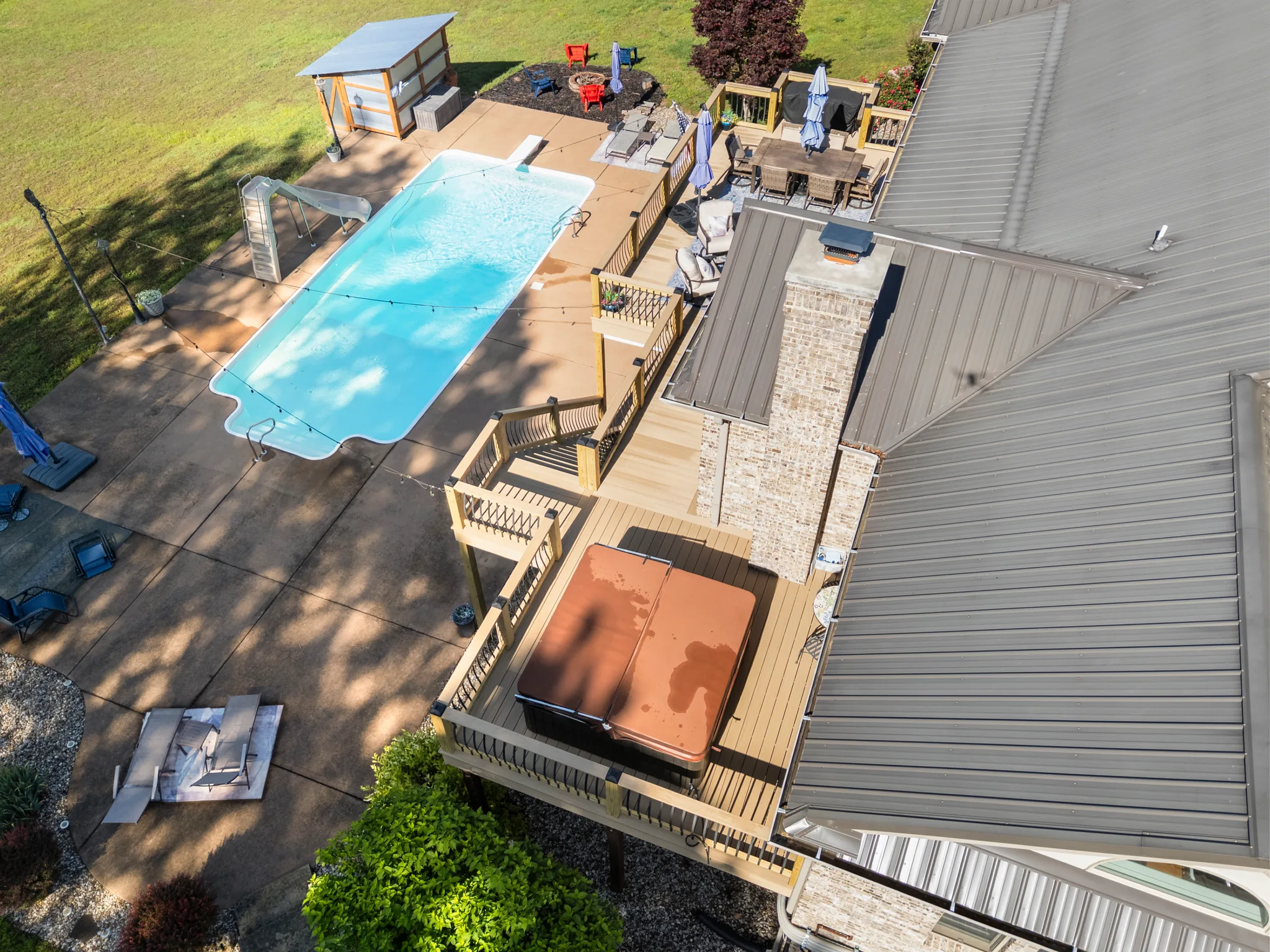 Aerial view of a multi-level composite deck with a hot tub and stone chimney beside a rectangular pool and concrete patio