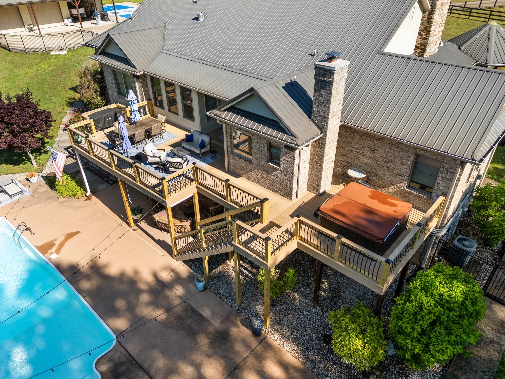 Aerial view of a multi-level wood deck along a brick home with a covered hot tub, dining set, umbrella and poolside concrete patio below
