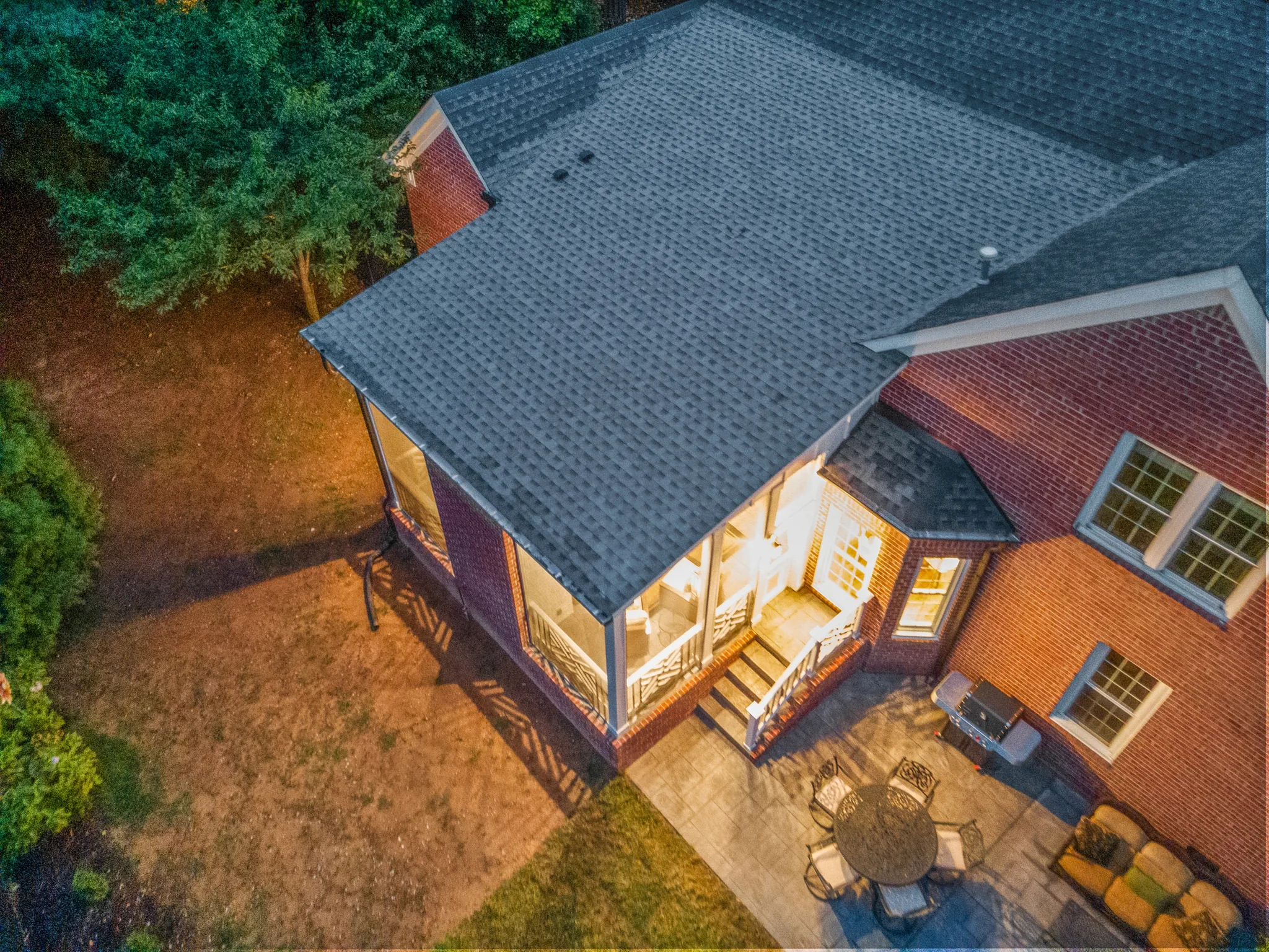Aerial view at dusk of a brick home with a shingled screened porch addition, lit interior, and a stamped patio with dining set and grill