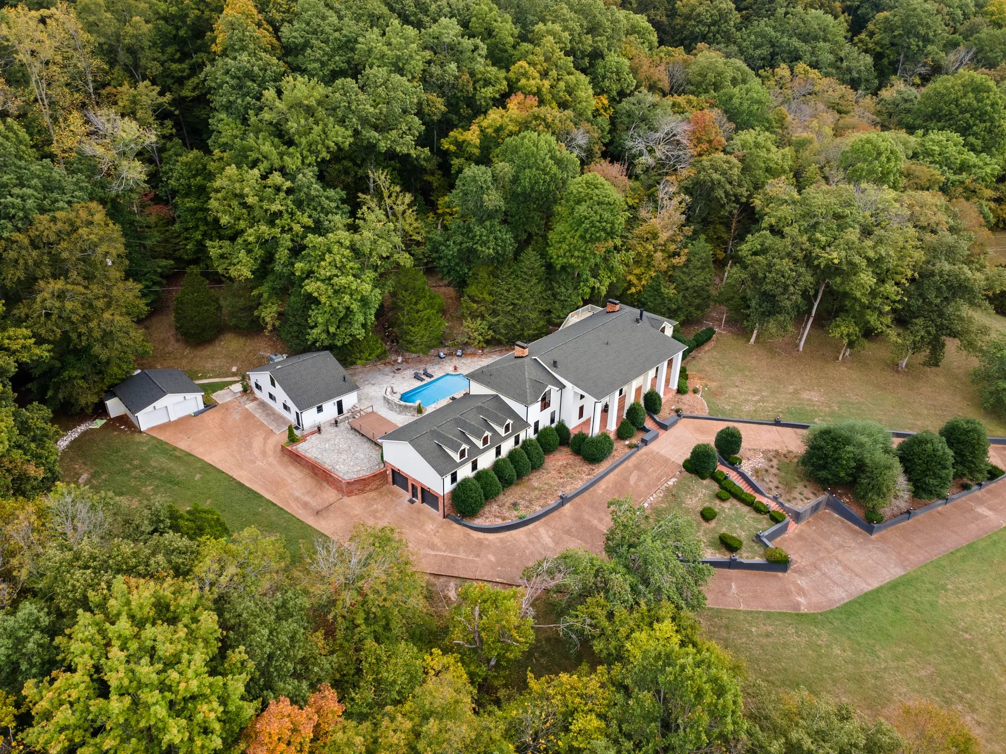 Aerial view of a white home with gray shingle roofs, a rectangular pool, detached garage and outbuilding, curving driveway and surrounding wooded hillside