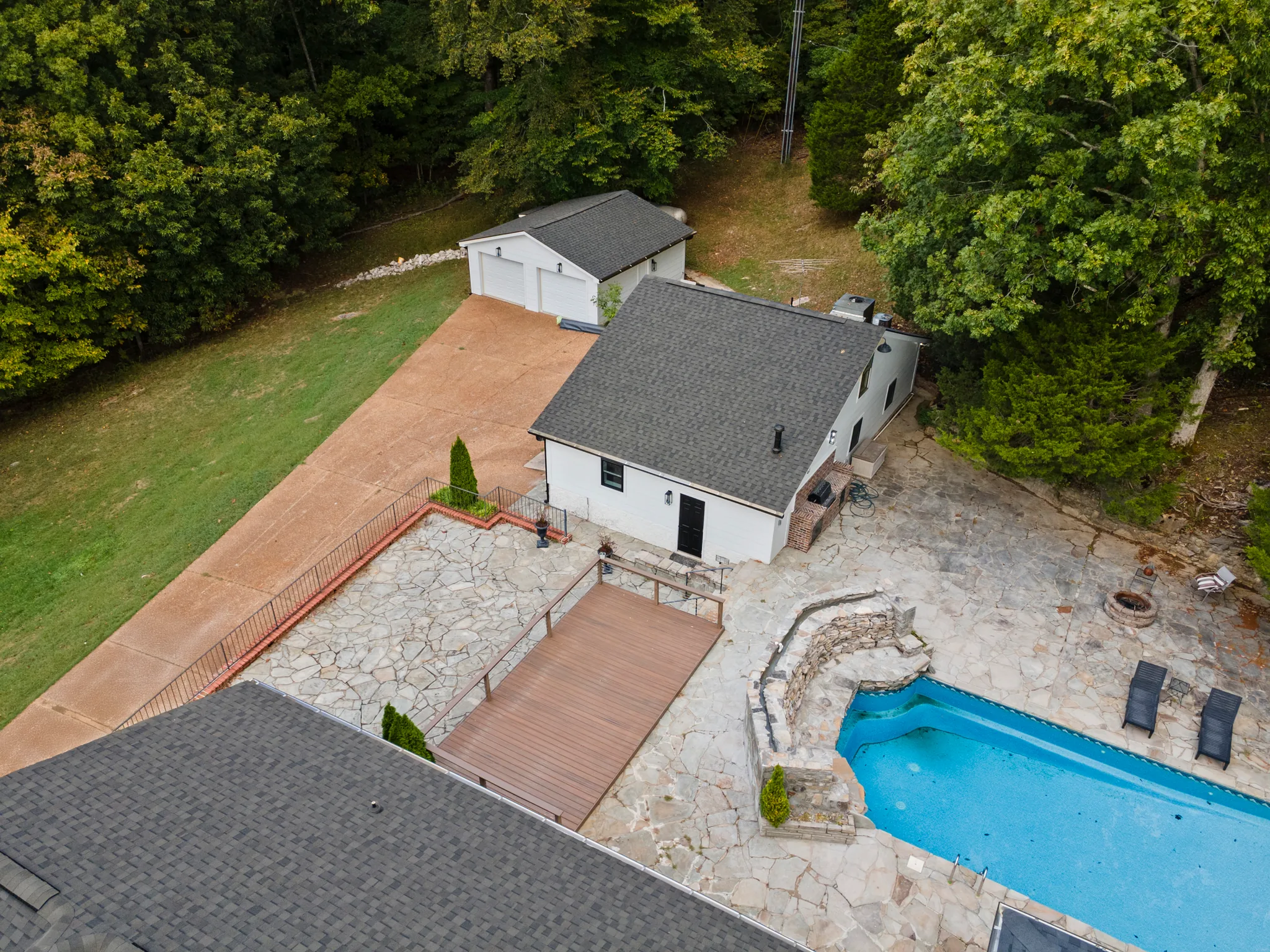 Aerial view of a white house and detached garage with a flagstone patio, composite deck platform, stone retaining wall and in-ground pool surrounded by woods