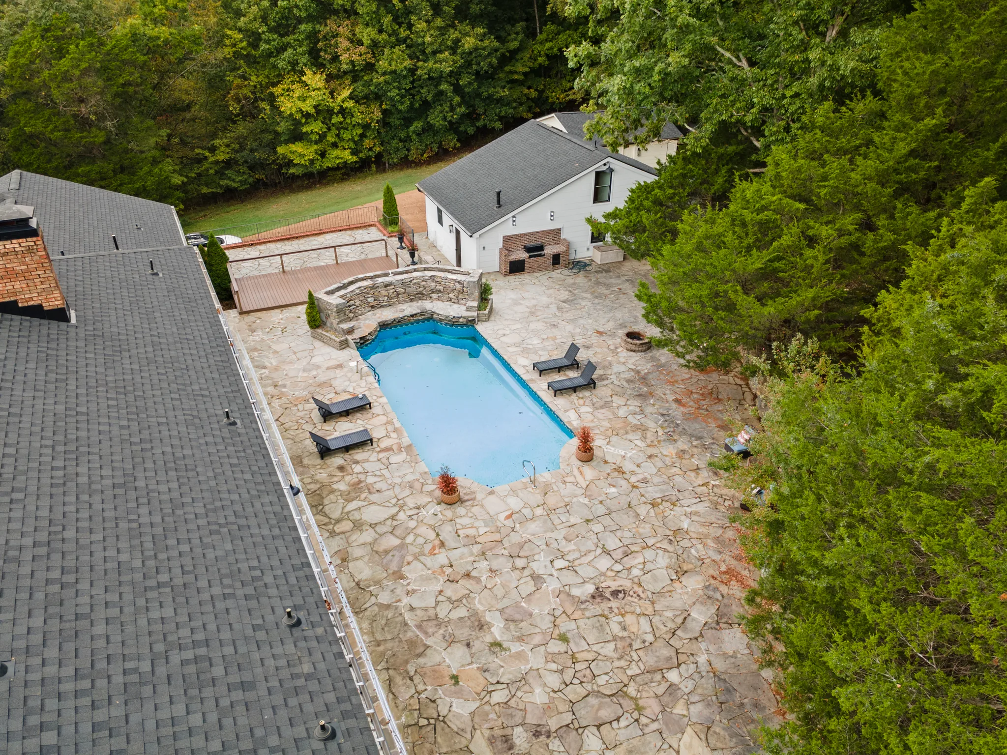 Aerial view of a rectangular pool surrounded by a wide flagstone patio, a stone retaining wall with spa, fire pit and lounge chairs between two homes