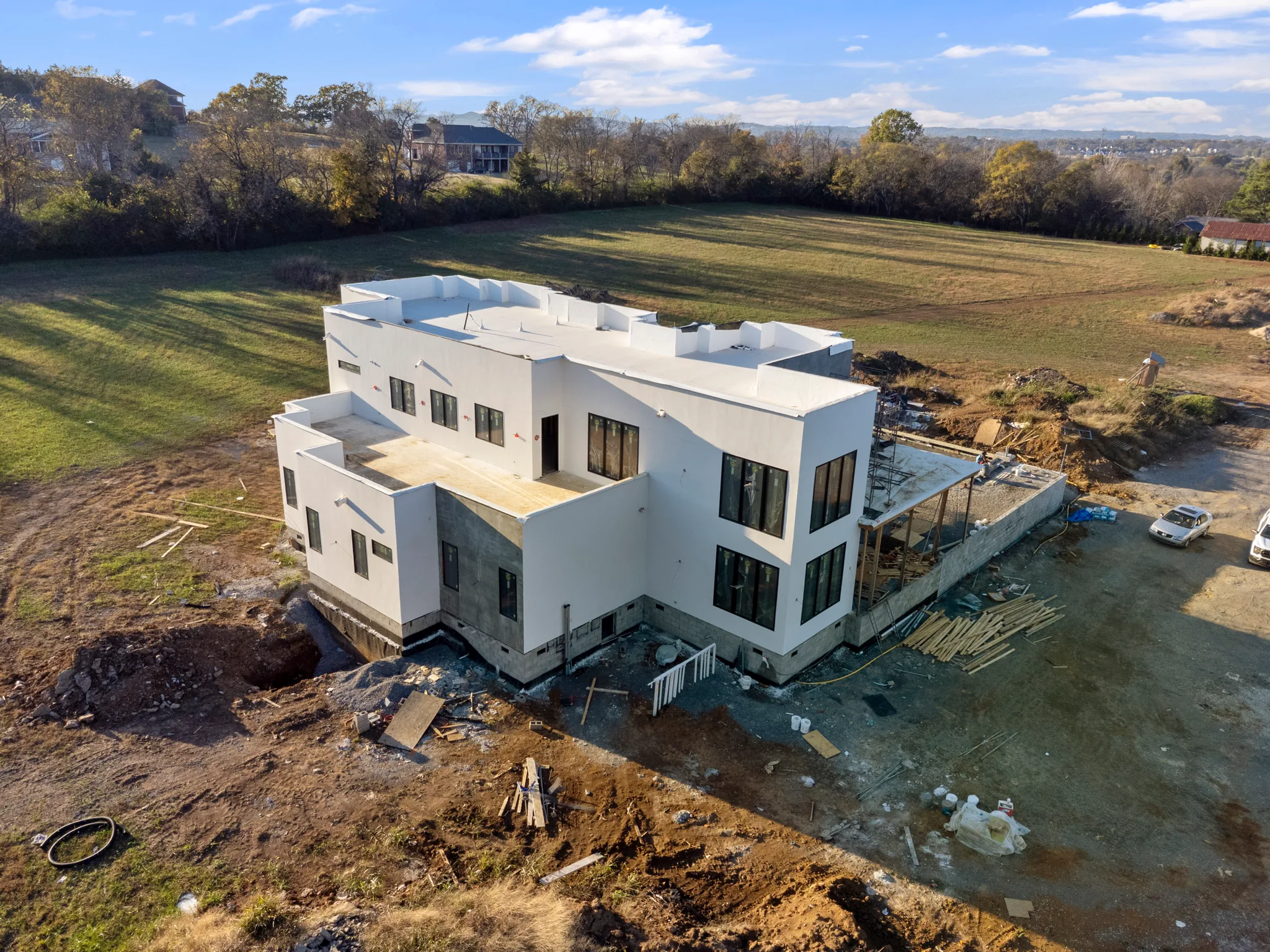 Aerial view of a two-story white flat-roof home under construction on a bare dirt site beside an open green field and trees