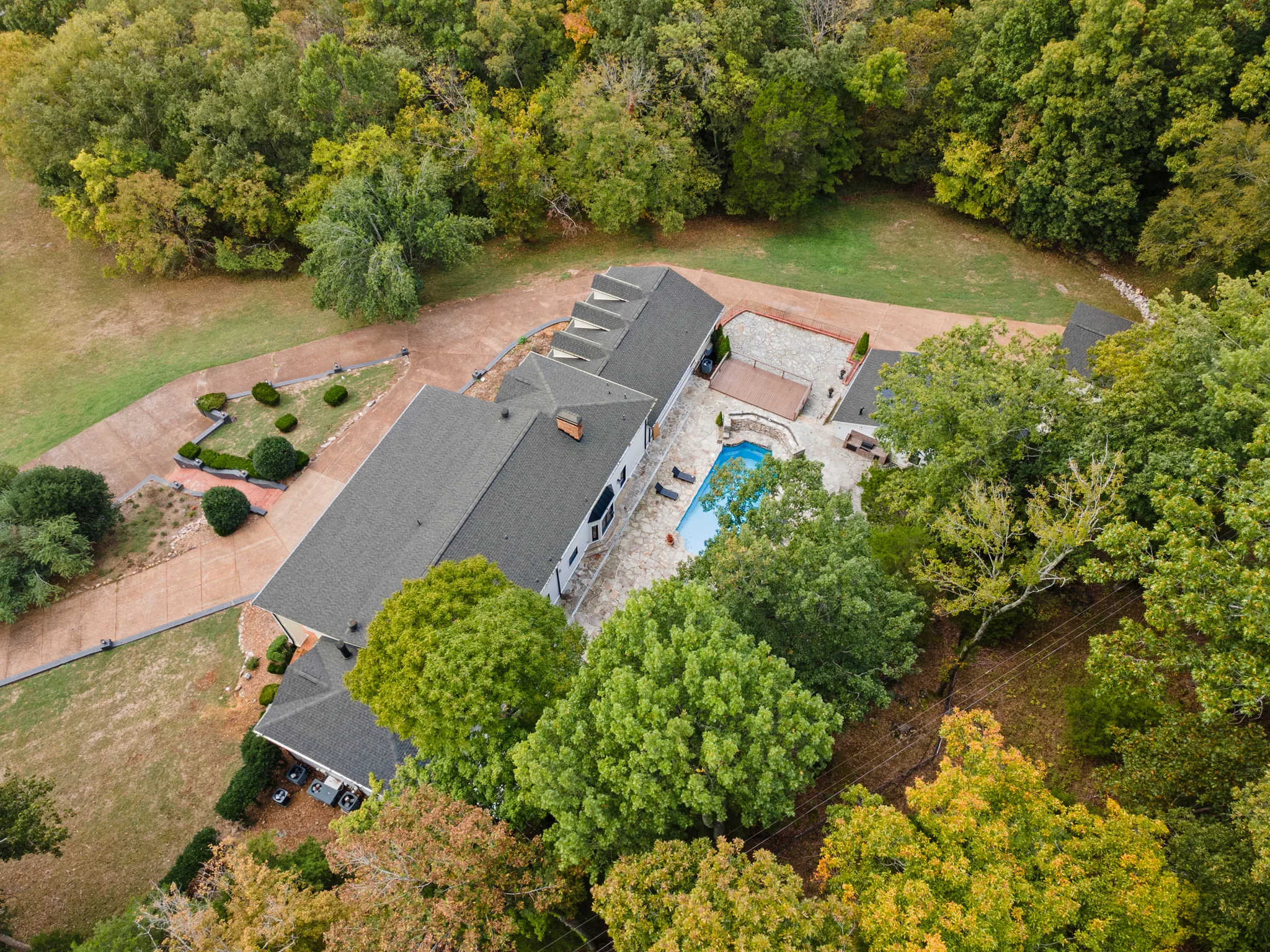 Aerial view of a long shingle-roofed home with a rectangular pool, stone patio, covered pavilion and curving drive surrounded by dense trees