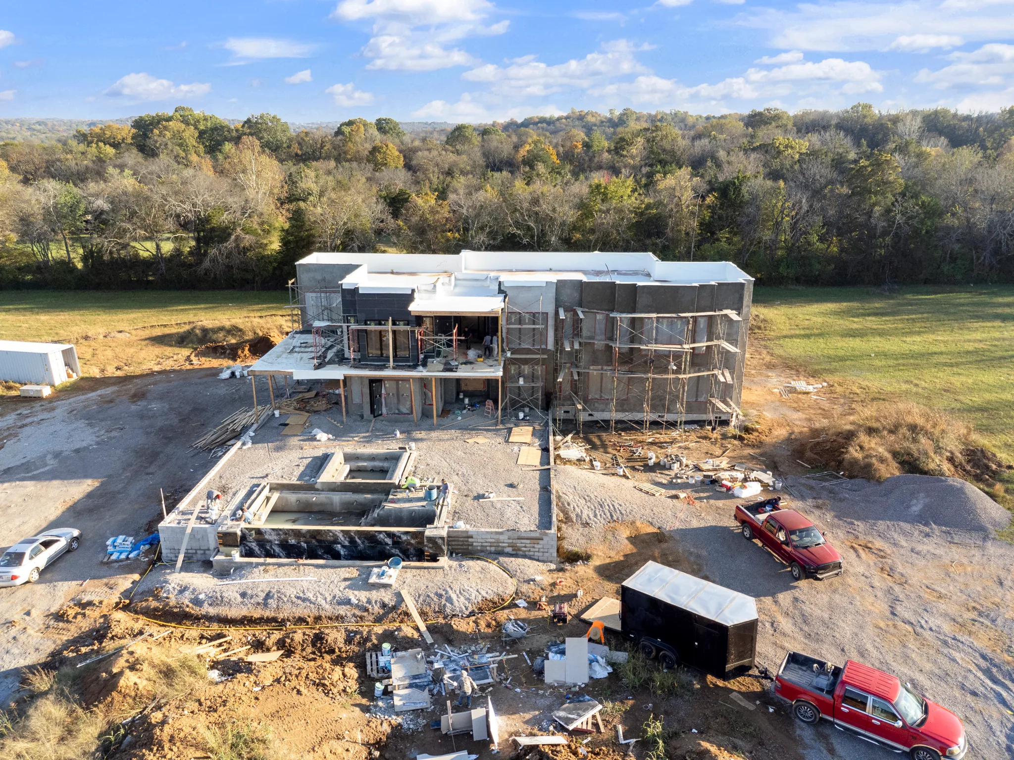 Aerial view of a large two-story home under construction with scaffolding, white flat roof sections and concrete pool shells on a graded gravel site bordered by trees