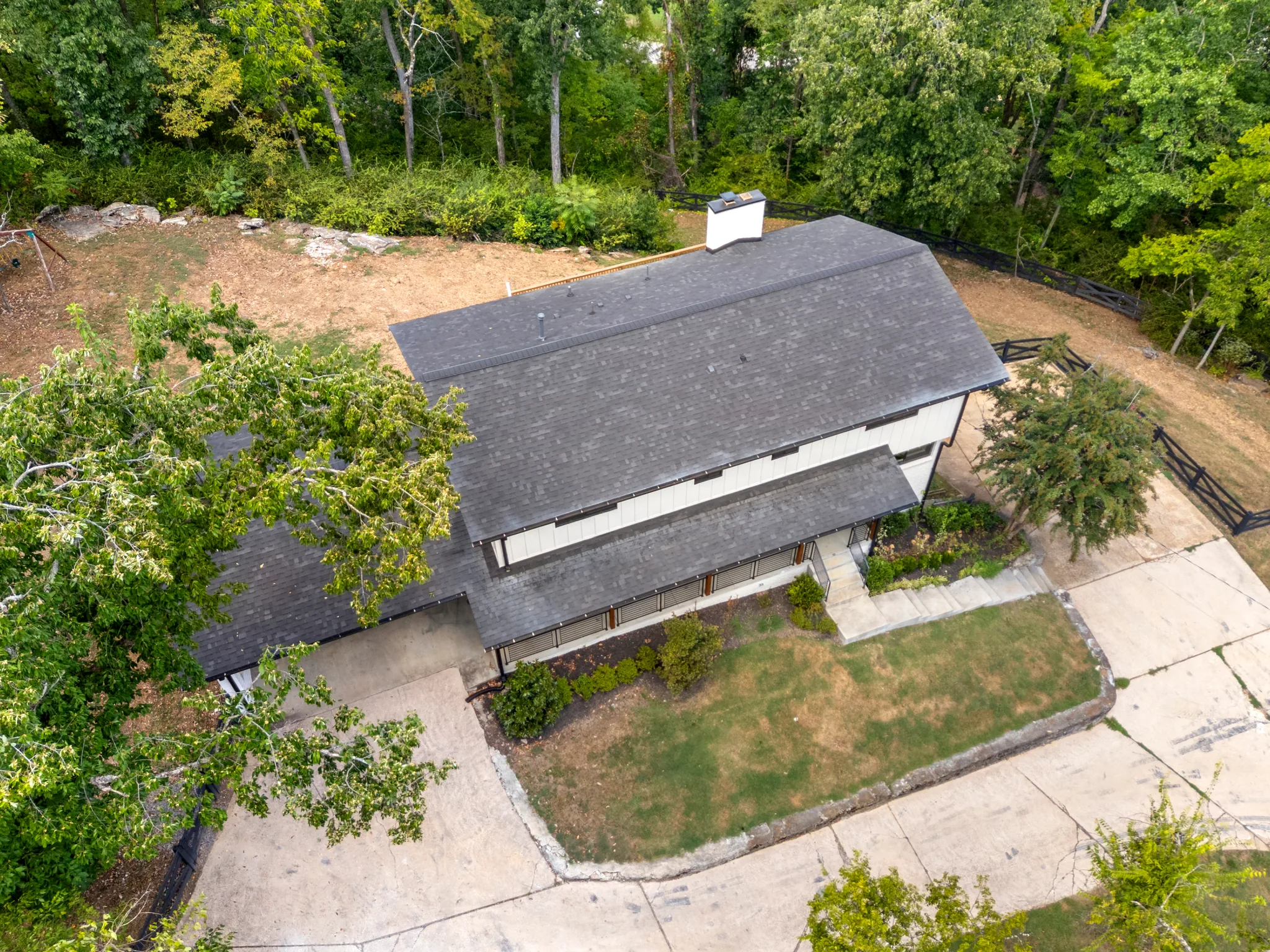 Aerial view of a modern home with a wide gray shingle roof, white vertical siding, a white chimney, concrete drive and wooded surroundings