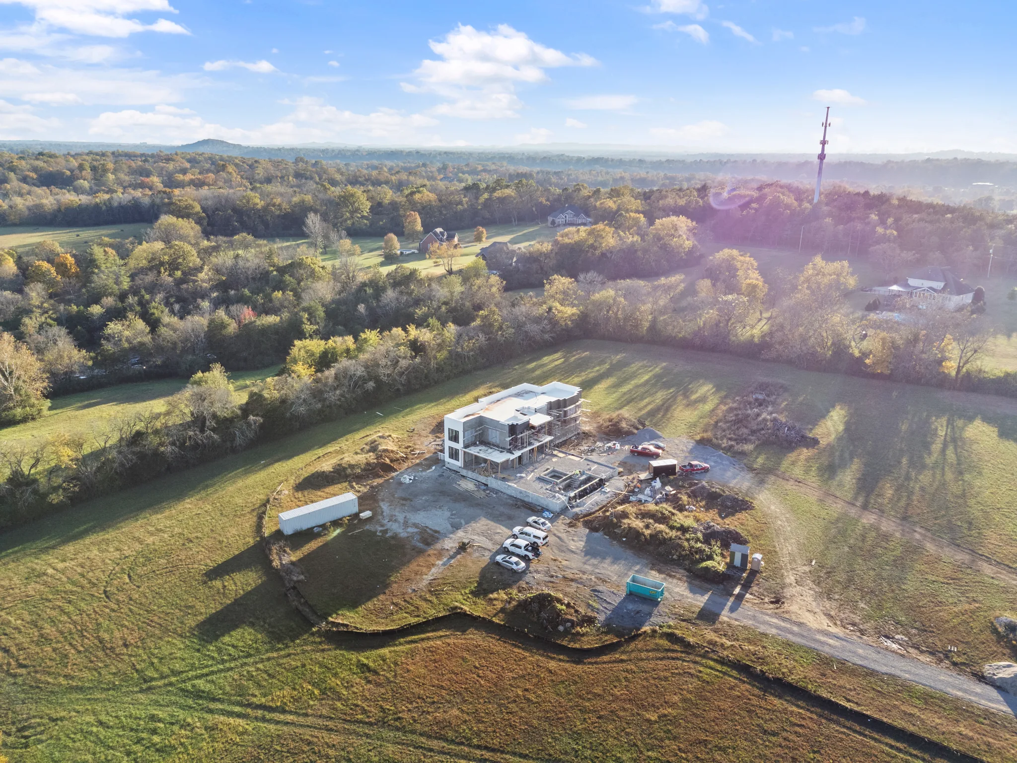 Aerial view of a white flat-roofed modern home under construction on a cleared pad surrounded by open fields and autumn woods