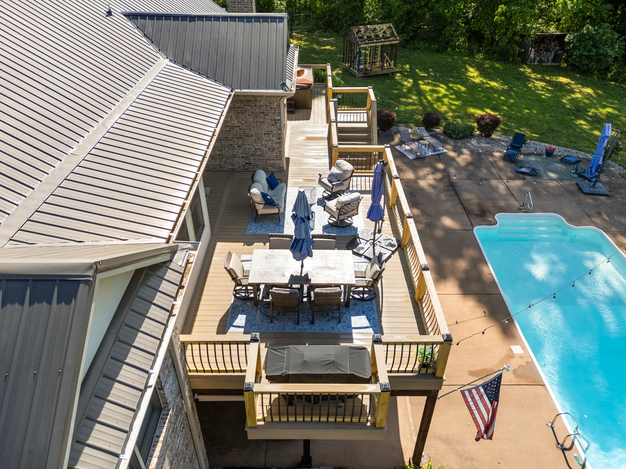 Aerial view of a multi-level composite deck along a brick and metal-roofed home, with dining set, lounge seating and umbrellas beside an in-ground pool