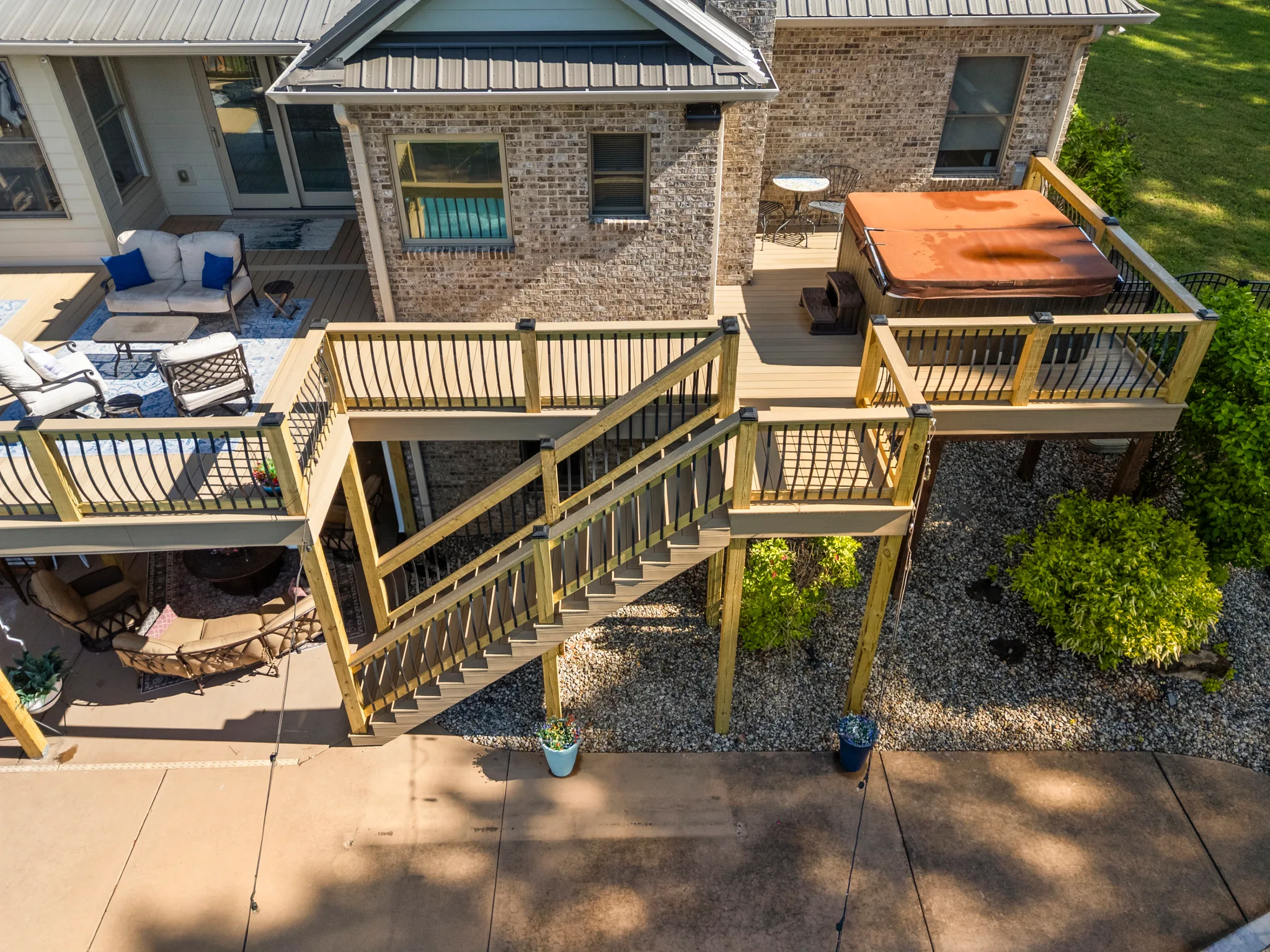 Aerial view of a multi-level composite deck on a brick home with wood framing, black metal balusters, a covered hot tub and a stair run to a concrete patio