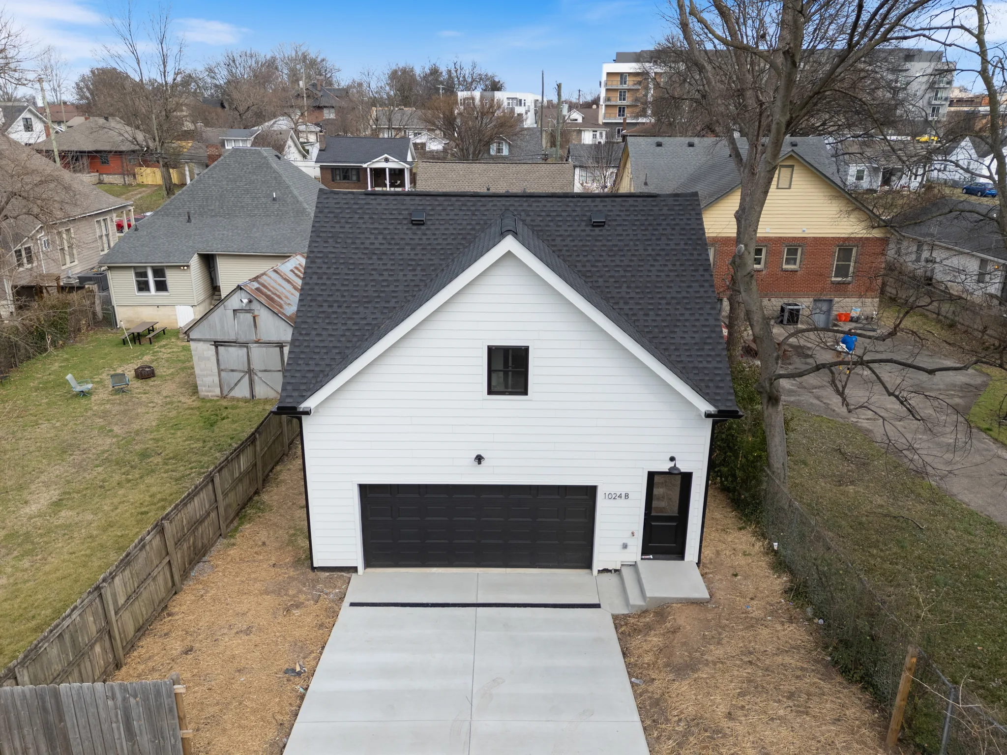 Aerial view of a white lap-sided detached garage with steep dark shingled gable roof, black overhead door and concrete driveway in a fenced city lot
