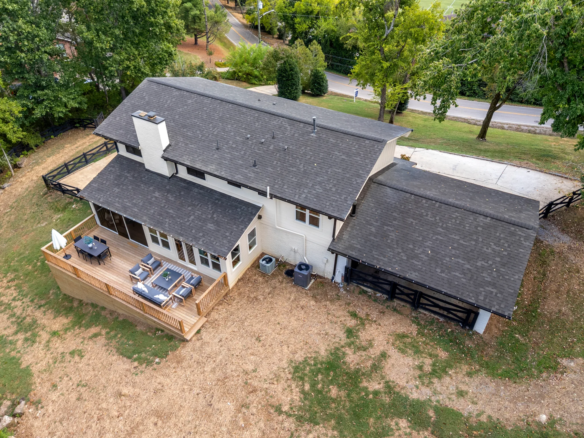 Aerial view of a renovated single-story painted brick home with dark shingle roofs, a wood rear deck with lounge seating, and an attached carport