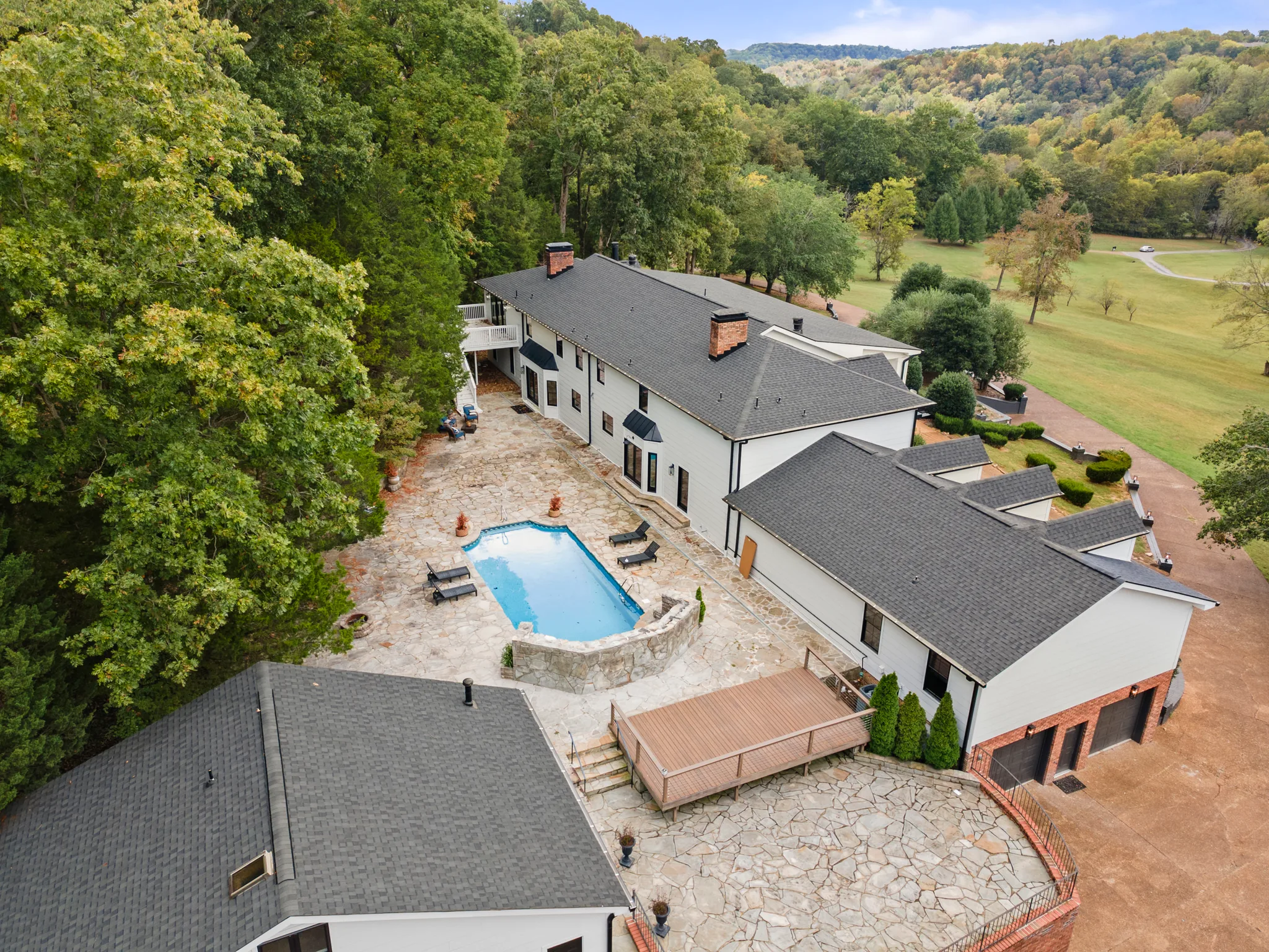 Aerial view of a large white house with dark shingle roofs wrapping an in-ground pool set in a wide flagstone patio beside woods
