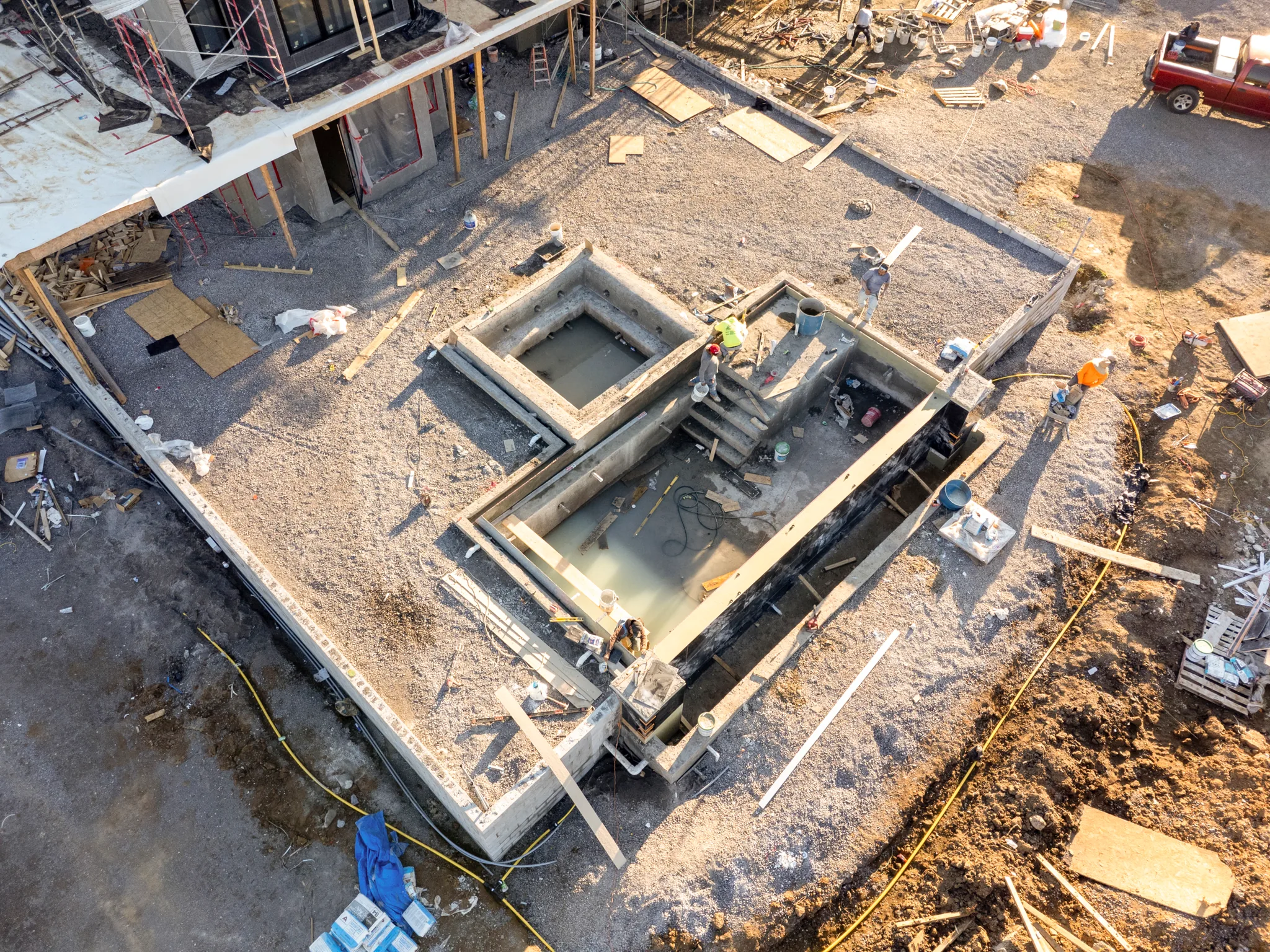 Aerial view of a concrete pool and spa shell under construction with gravel deck area, forms, crews and a building frame nearby