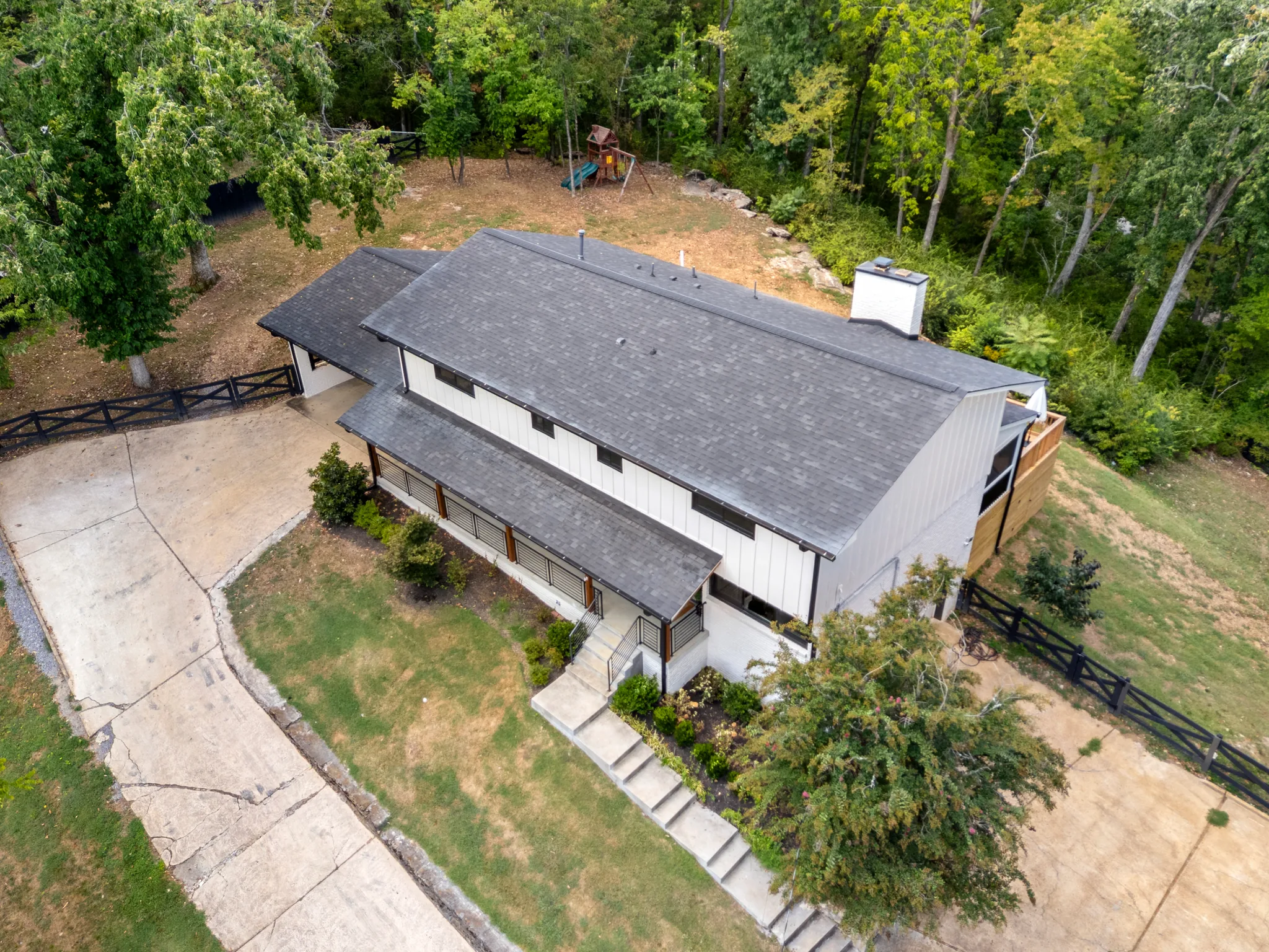 Aerial view of a long single-story home with white board and batten siding, dark shingle roof, covered porch, concrete drive and black fencing
