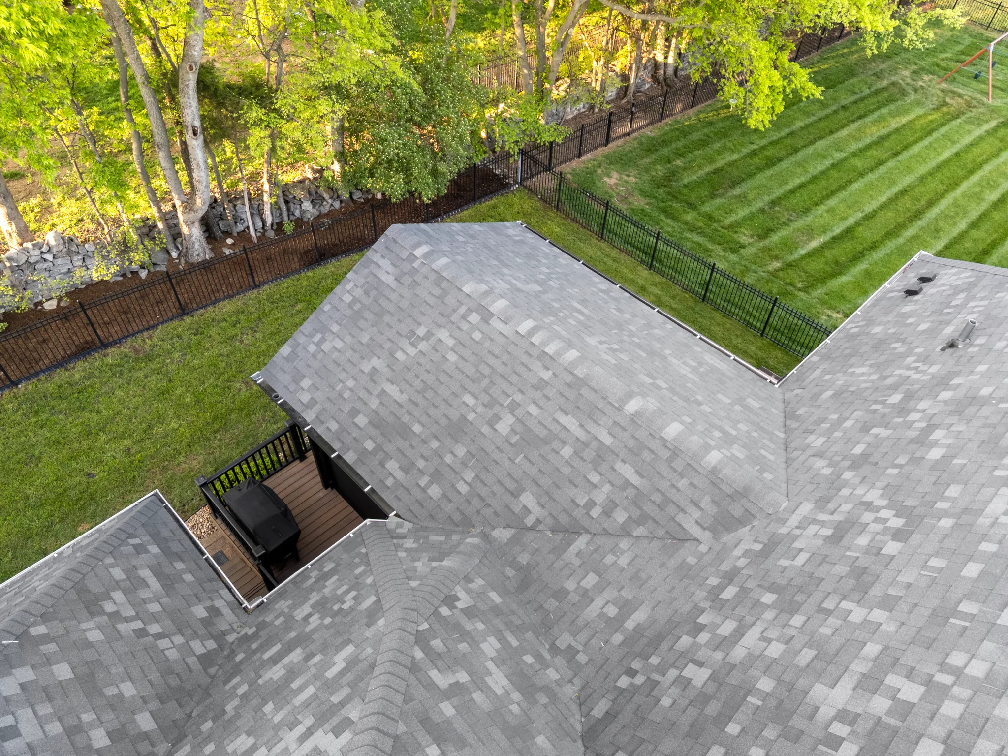 Aerial view of gray shingle roof gables with a small composite grill deck and black railing beside a fenced lawn and woods