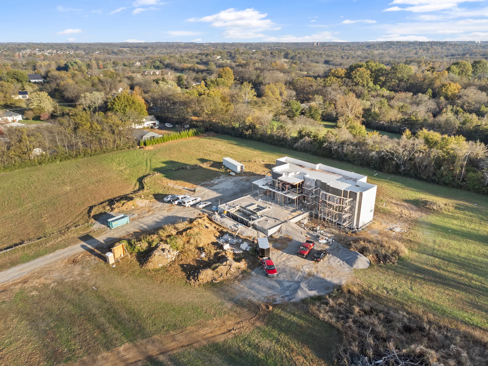 Aerial view of a large flat-roofed home under construction with white walls and scaffolding, surrounded by gravel drive, pasture and autumn trees