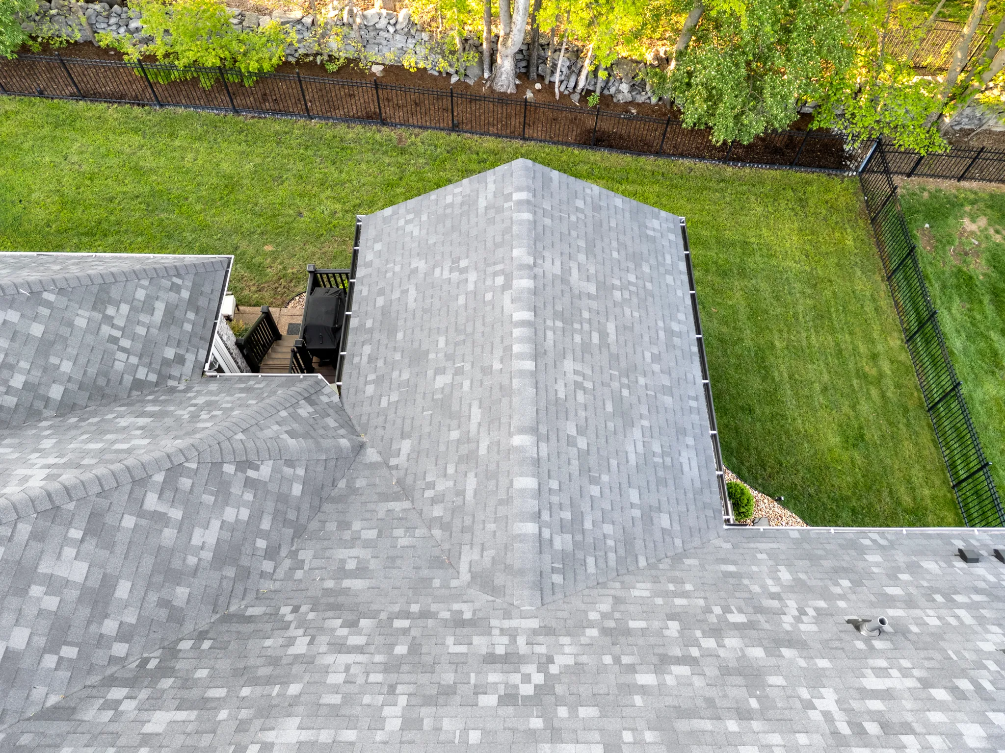 Aerial view of a gray architectural shingle roof over intersecting gables, with a small deck, grill and fenced green lawn below