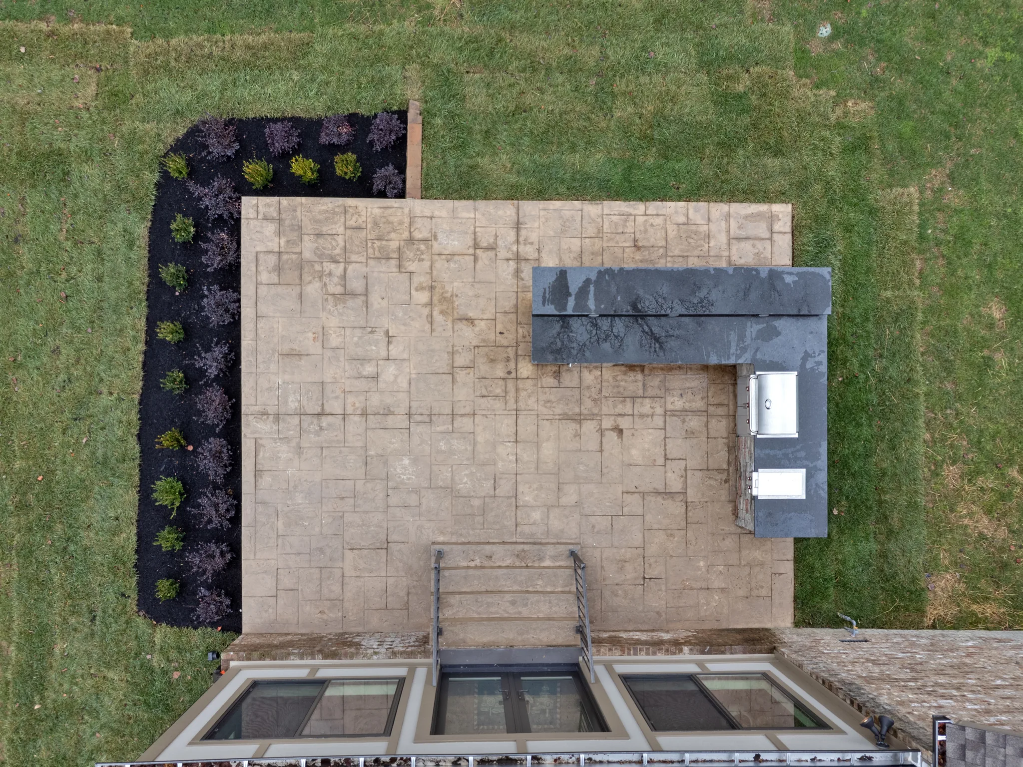 Aerial view of a stamped concrete patio with an L-shaped stone grill island, dark counters, mulched planting bed and lawn