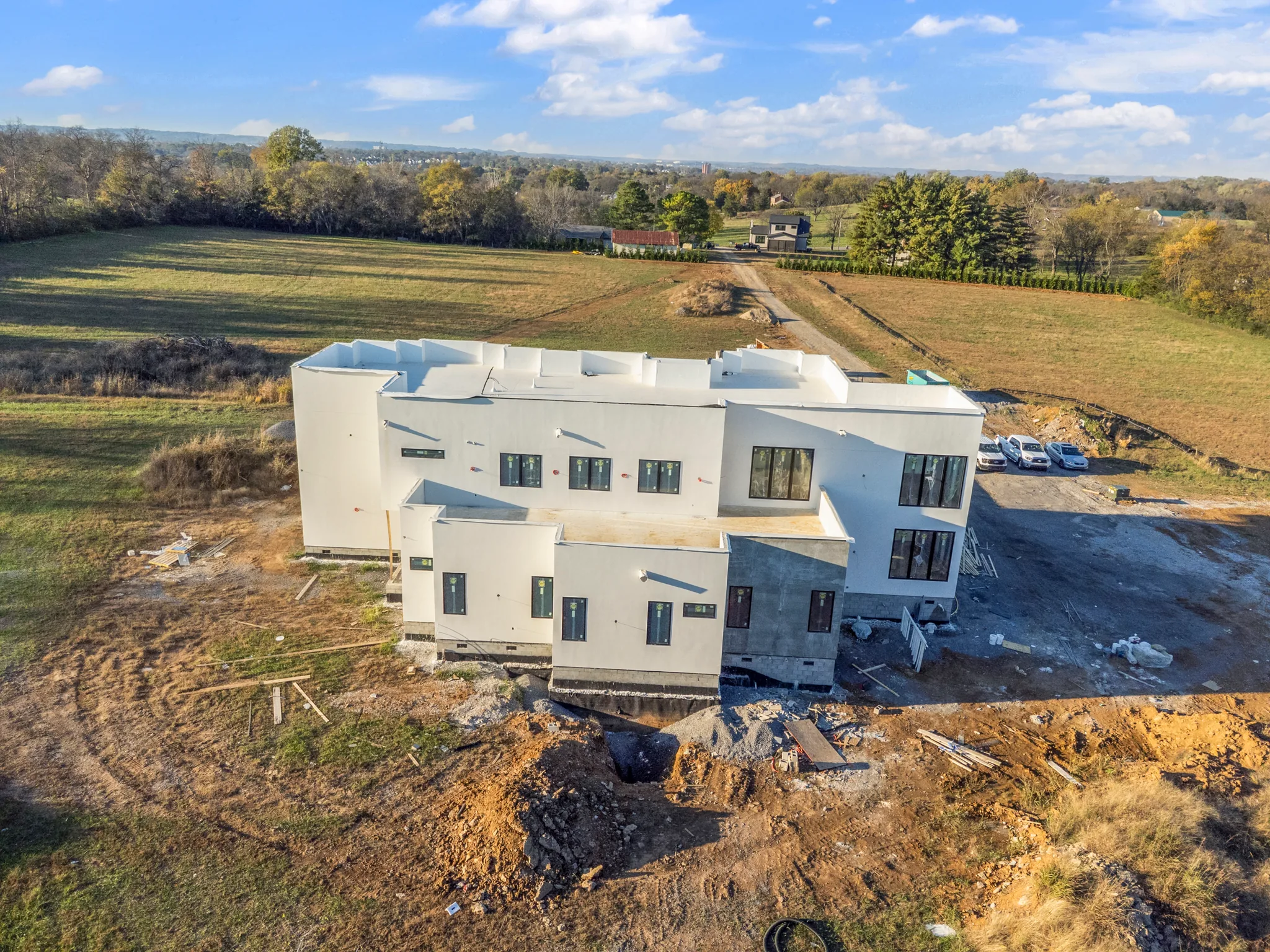 Aerial view of a large white flat-roof stucco home under construction on open farmland, with dark windows, graded dirt and parked cars