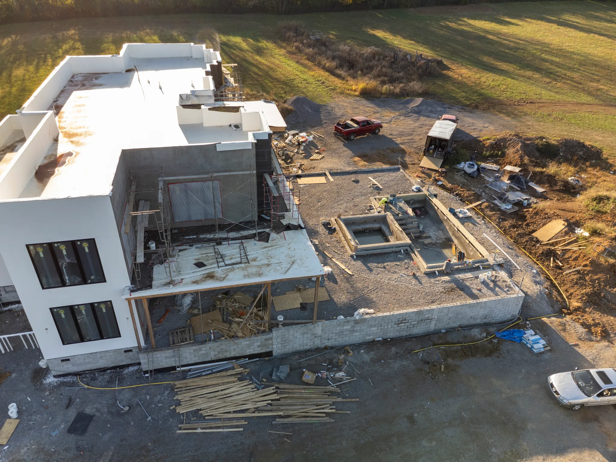 Aerial view of a white flat-roof stucco home under construction with block retaining wall, concrete pool forms, lumber piles and gravel work area