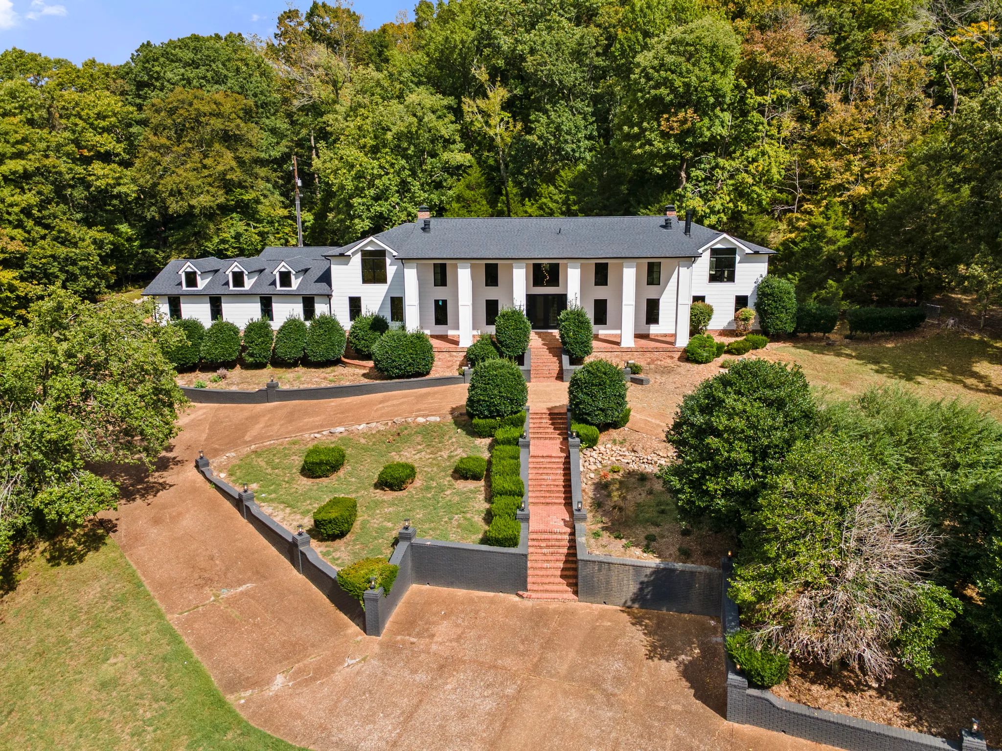 Aerial view of a long white two-story home with black shingle roof, tall front columns, dormer wing, and a brick stair path rising through terraced landscaping