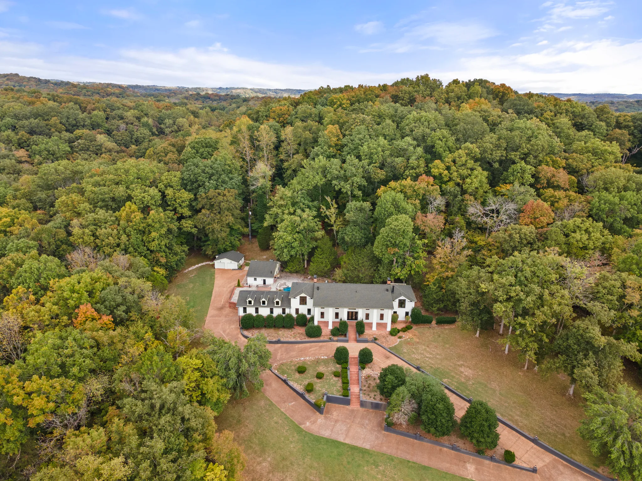 Aerial view of a long white home with dark shingled roof, dormers and columned entry, set on a lawn surrounded by wooded hills