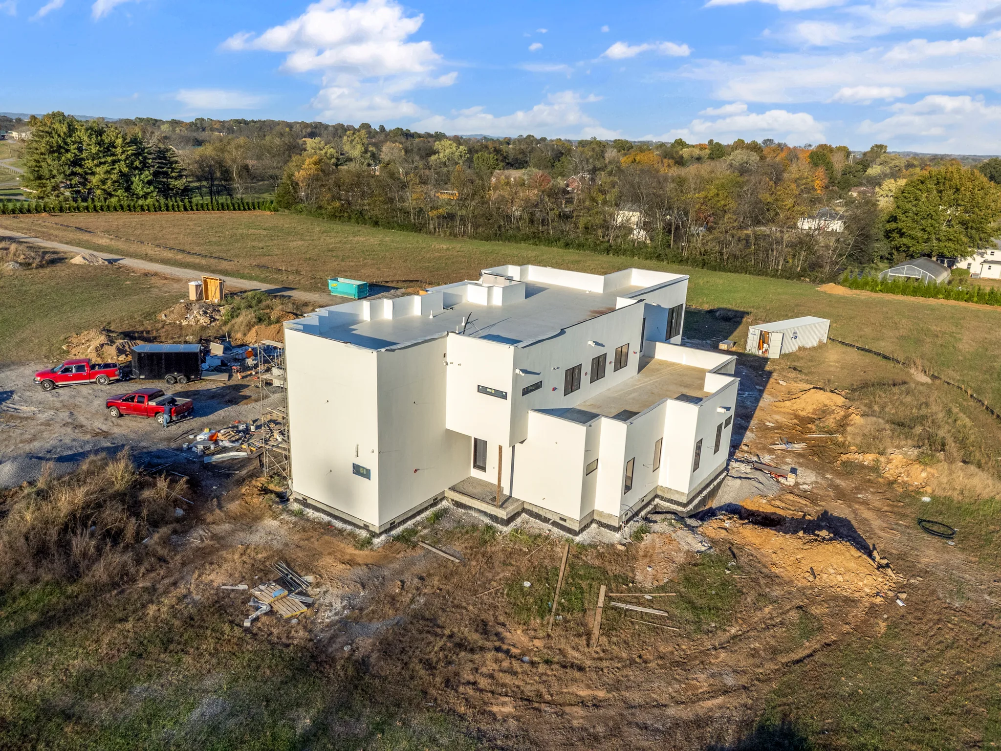 Aerial view of a white stucco flat-roof home under construction on a cleared lot with pickup trucks, dumpster and fields beyond