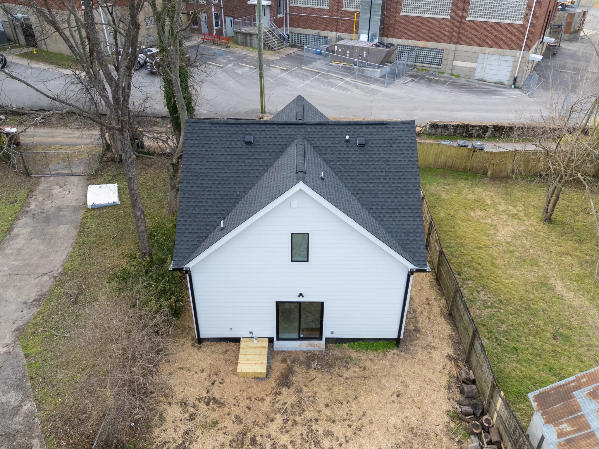 Aerial view of a white lap-sided home with dark shingle gable roof, sliding glass door, small wood landing and fenced dirt yard