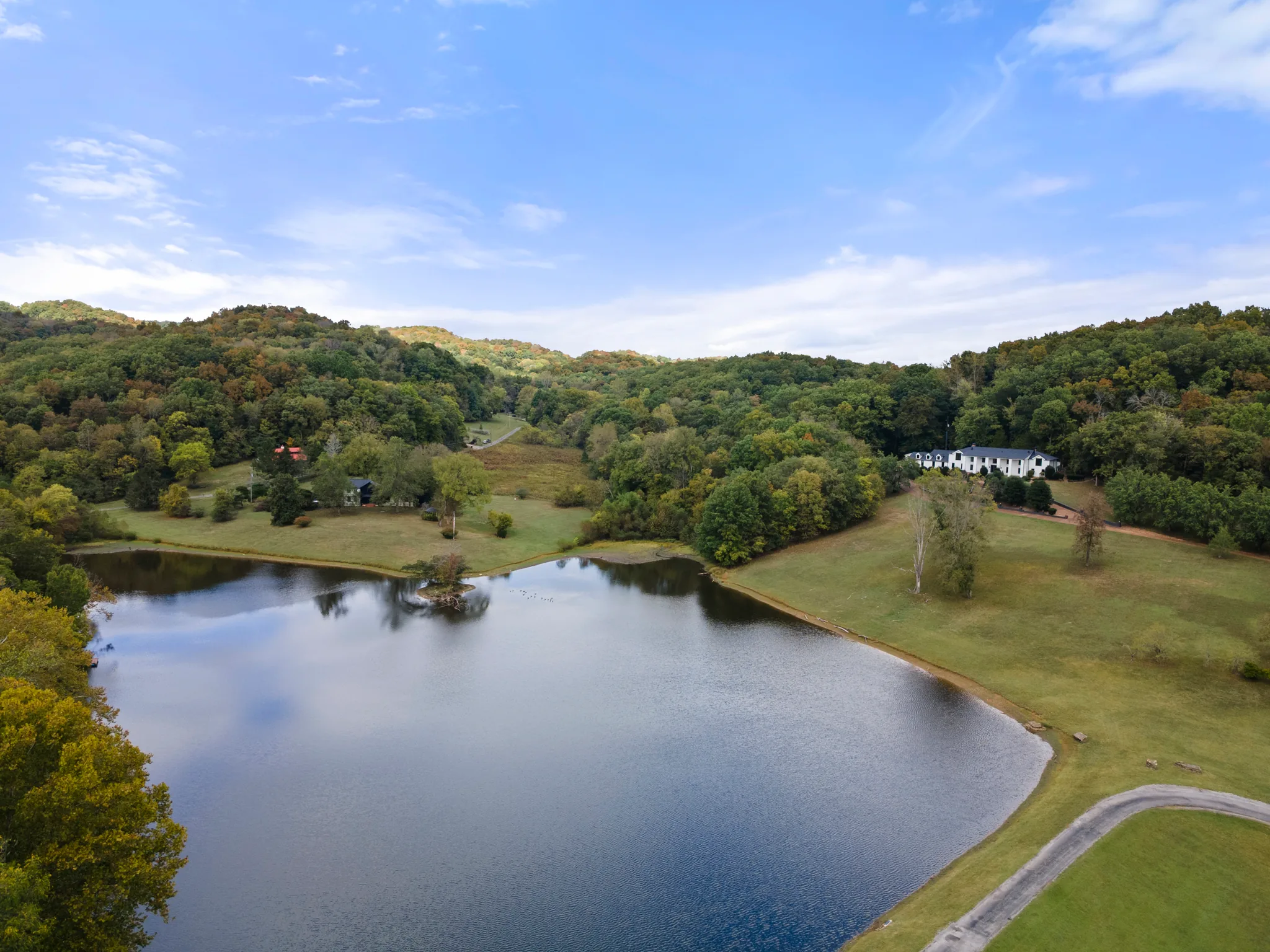 Aerial view of a white two-story home with dark metal roof set on a wooded hillside above a large lake and mowed lawn