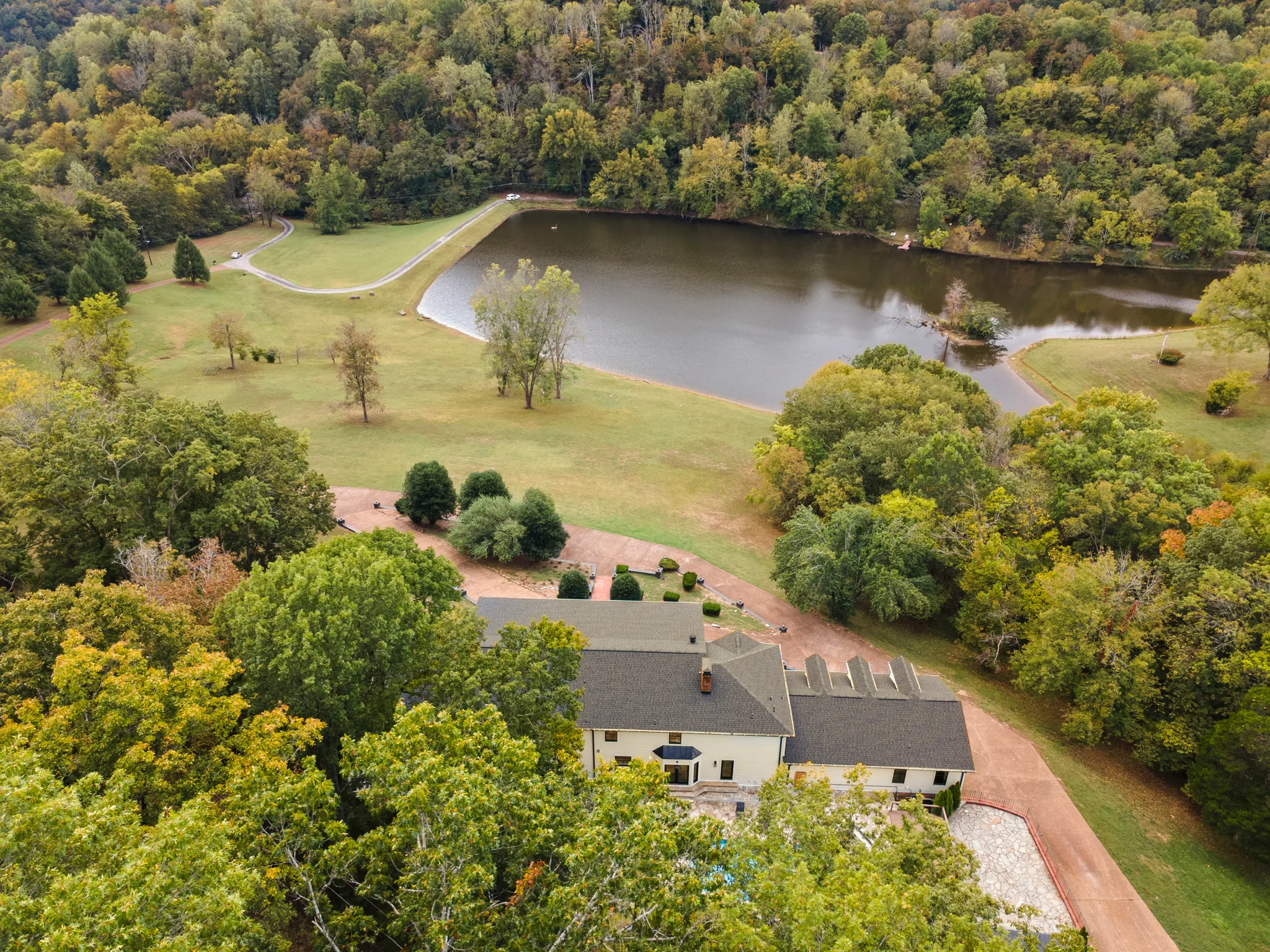 Aerial view of a white home with gray shingle roofs set among mature trees, with a mowed lawn, gravel drive and pond beyond