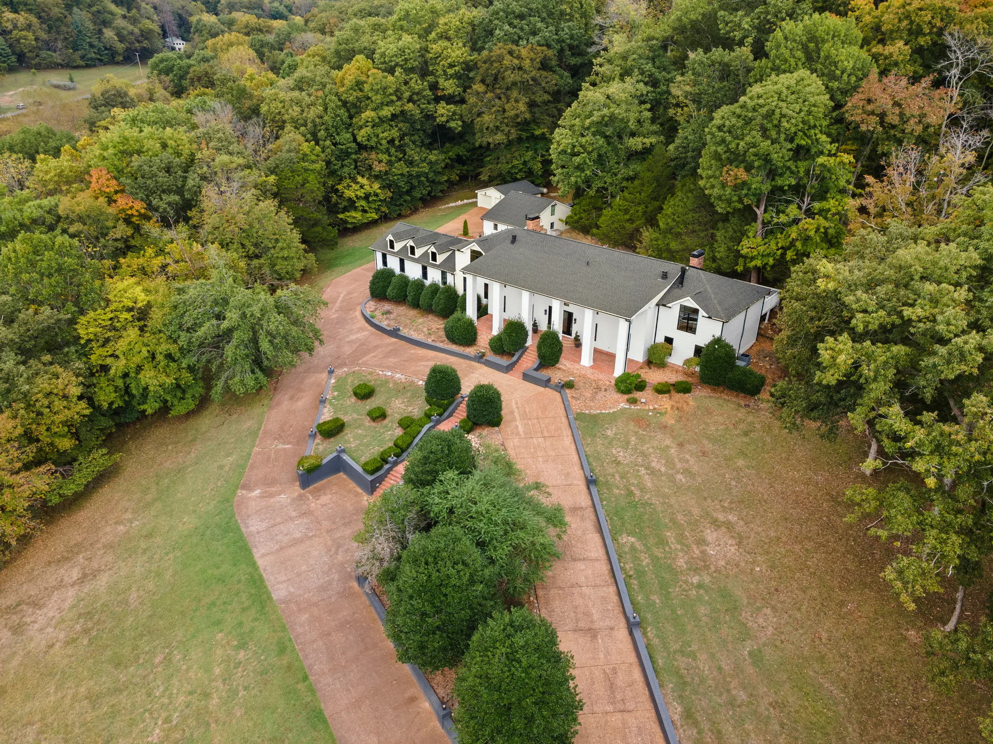 Aerial view of a long white house with dark shingle roof, dormers and columned porch, set behind a wide circular drive and trimmed shrubs