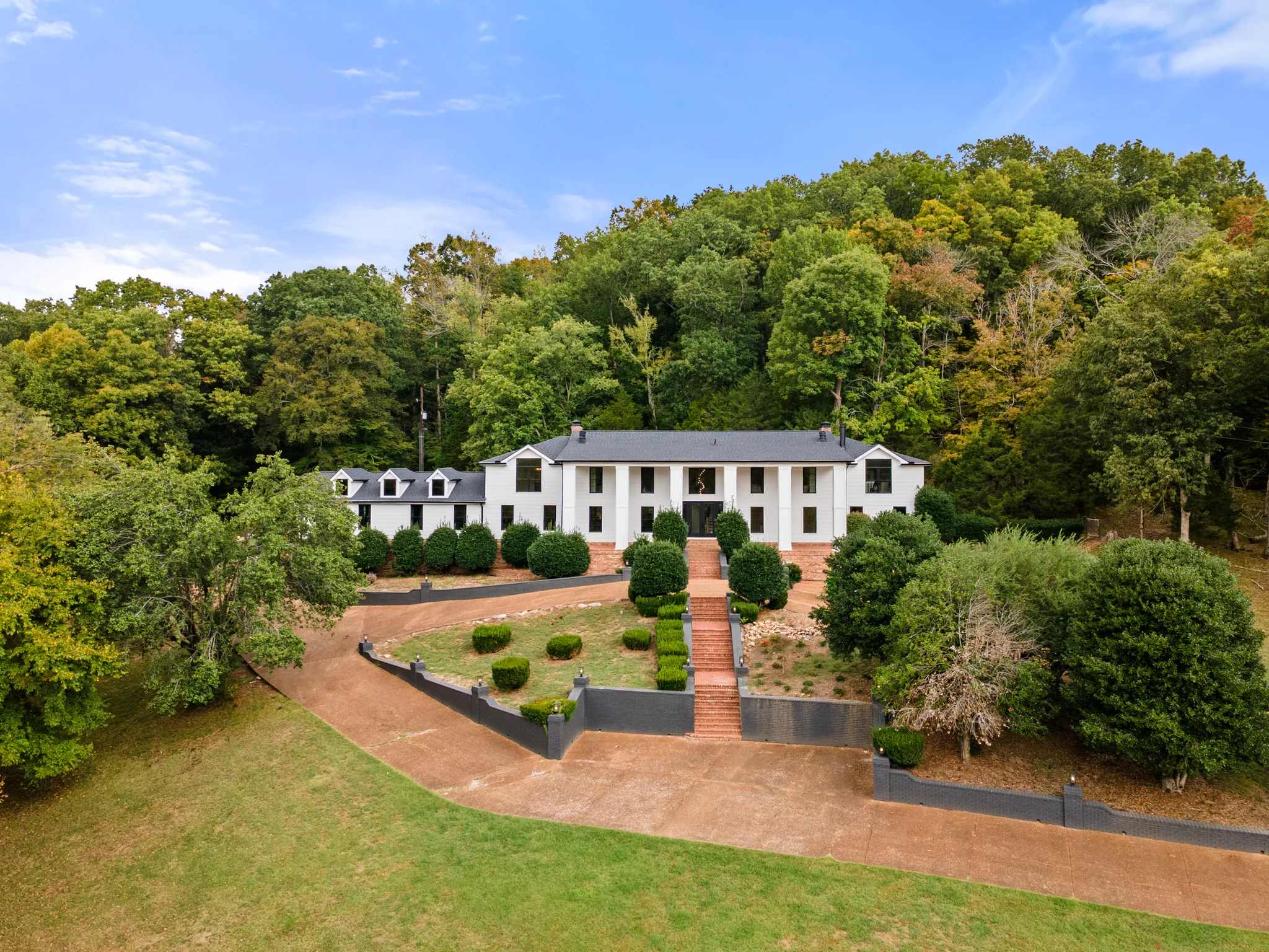 Aerial view of a long white two-story home with black shutters, columned center entry, dormer wing, and brick stairs rising through terraced landscaping to the front door