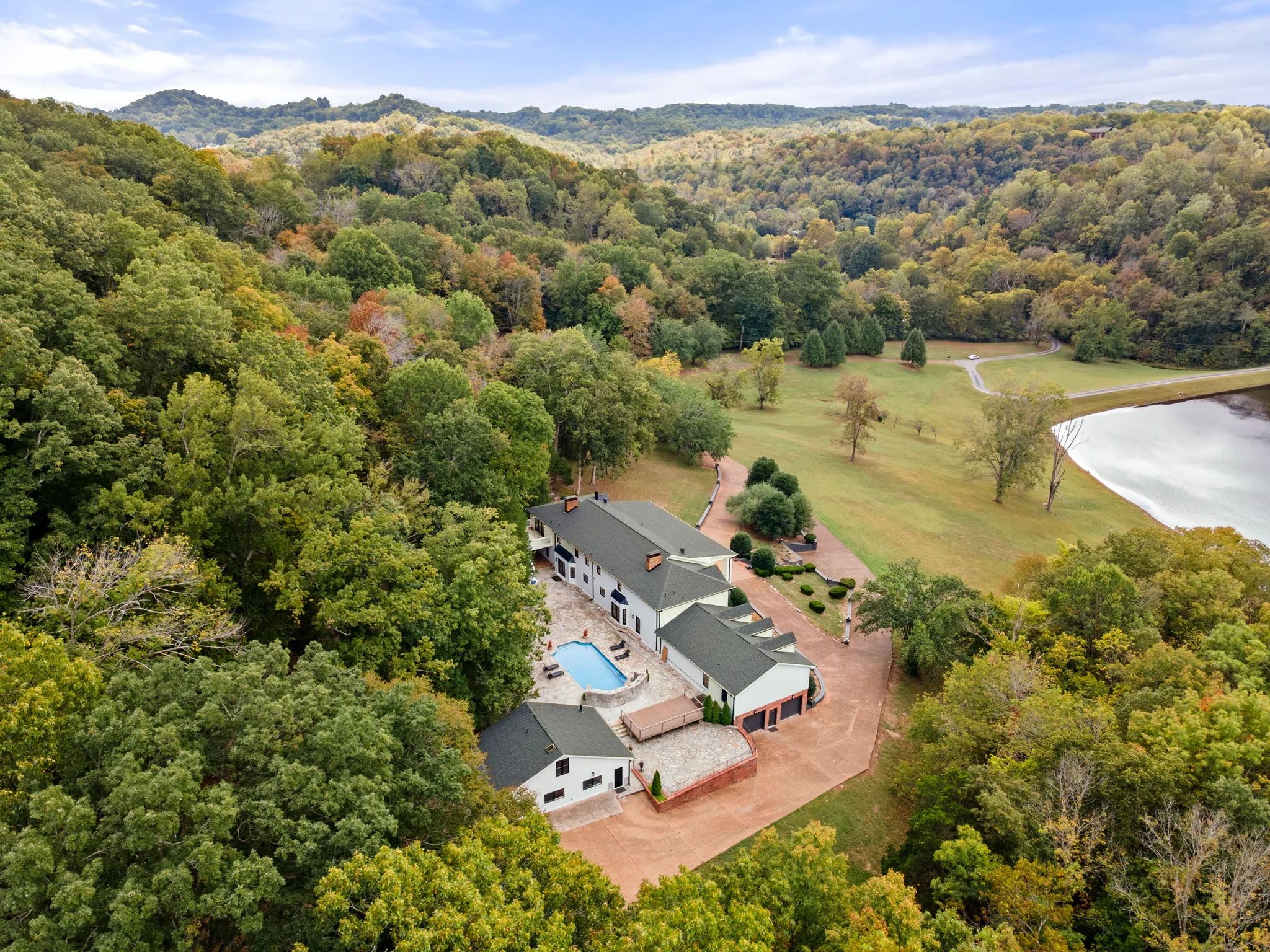 Aerial view of a long white home with dark gable roofs, pool and paved courtyard set in a wooded valley beside a lake and open lawn