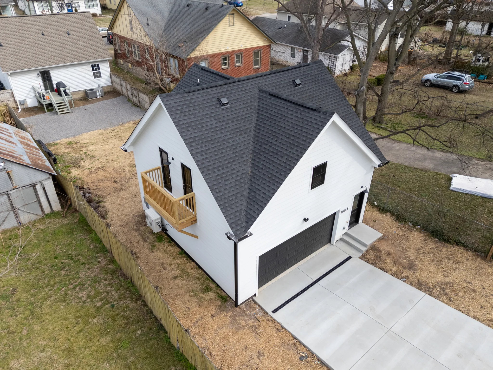 Aerial view of a compact white home with dark shingled gable roof, attached garage, concrete drive, and a small wood balcony