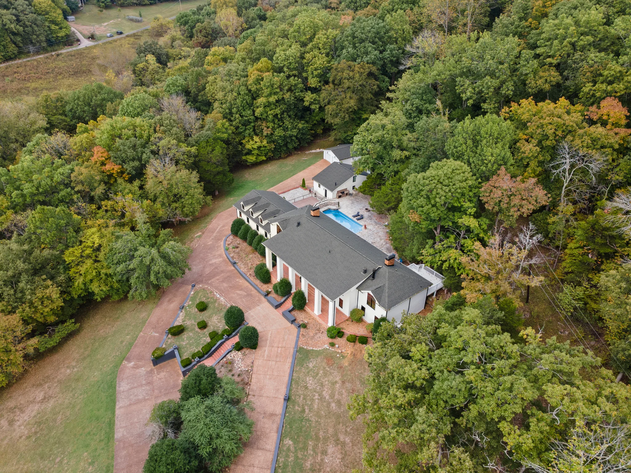 Aerial view of a white house with dark shingle roof, brick drive and walkways, and a rectangular pool surrounded by mature woods