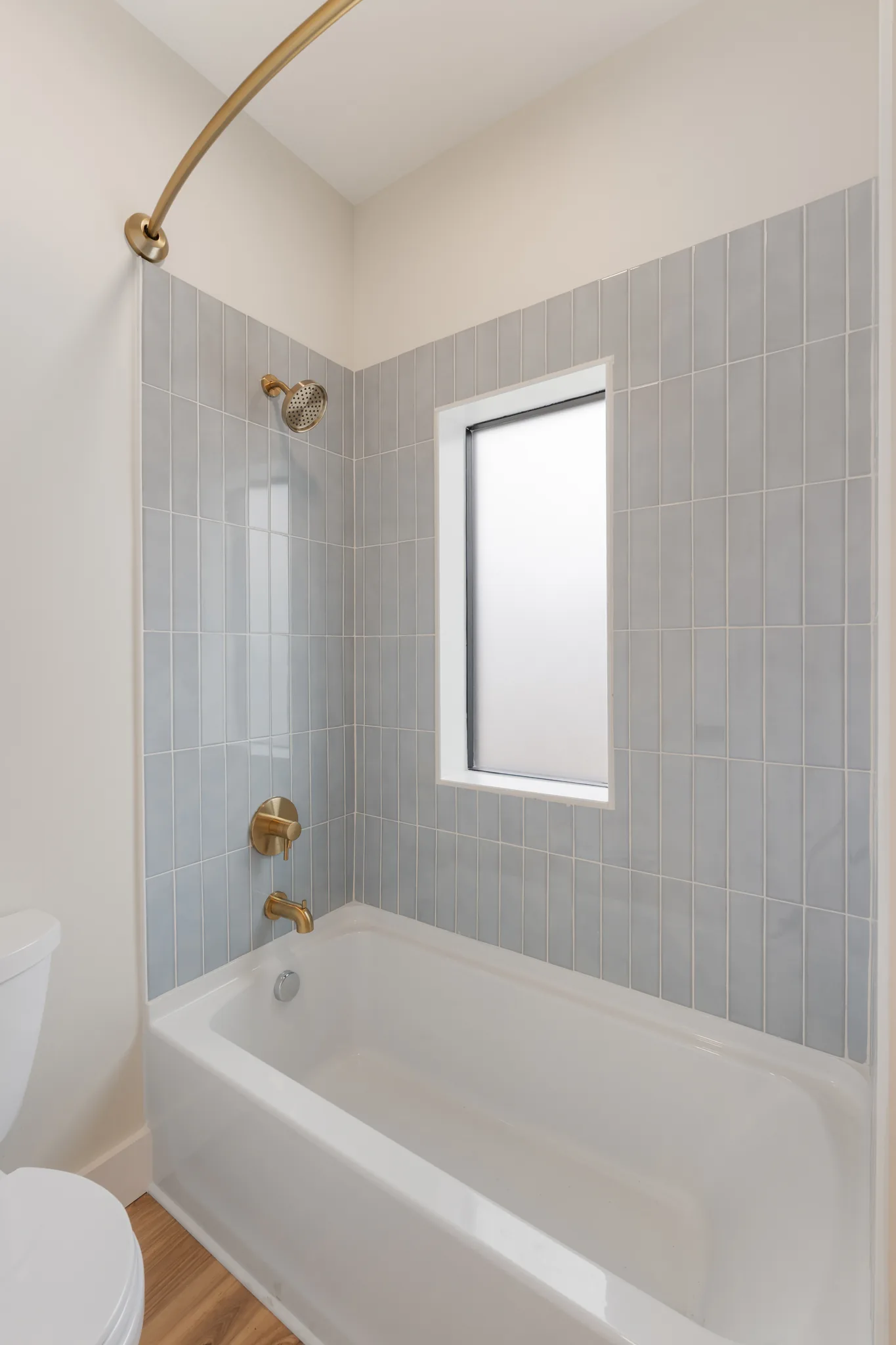 White alcove bathtub surrounded by light blue vertical stacked tile with brass shower fittings, a curved rod and a frosted window