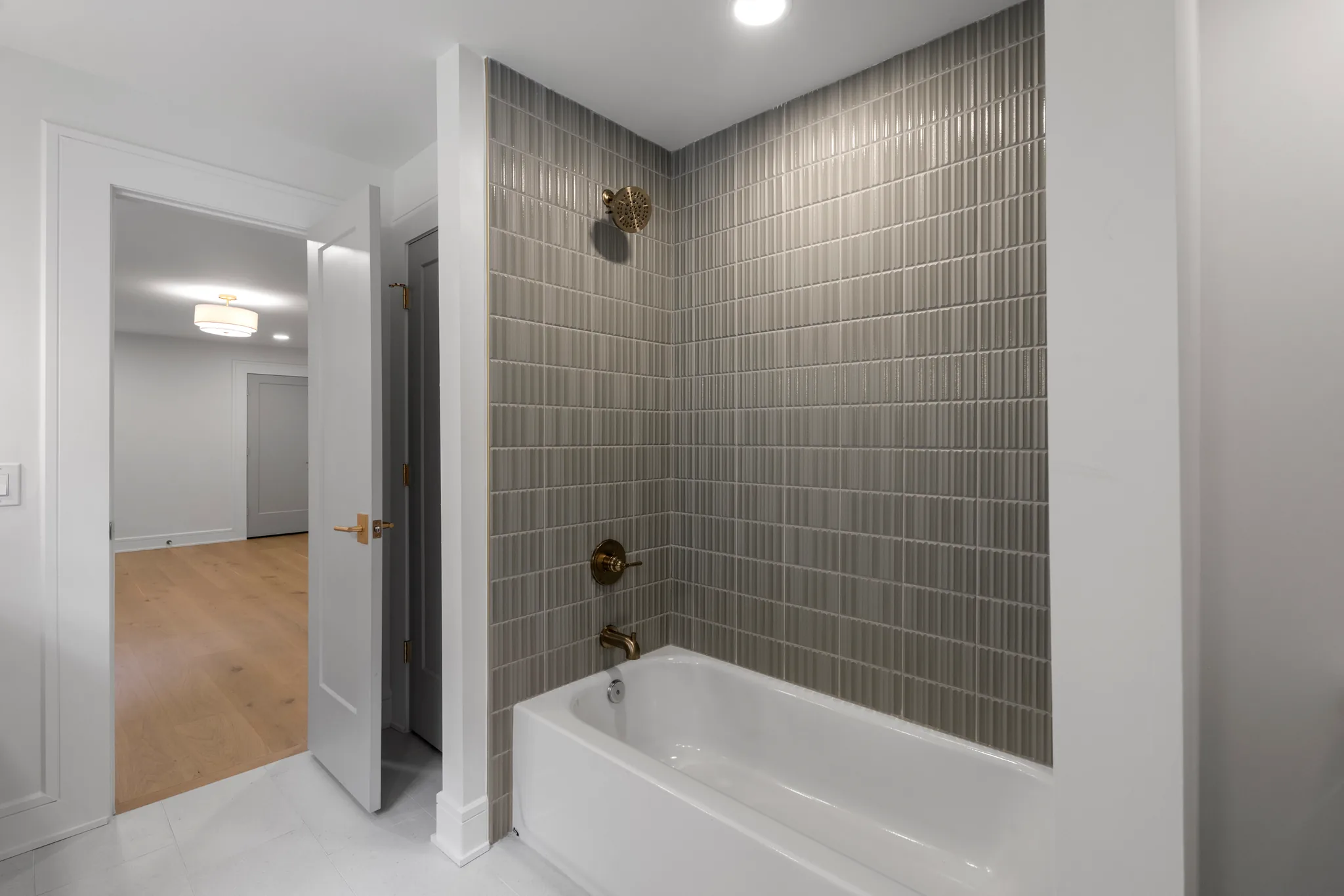 Bathroom alcove tub surrounded by gray vertical stacked tile with brass shower head and tub filler, open door to hallway beyond