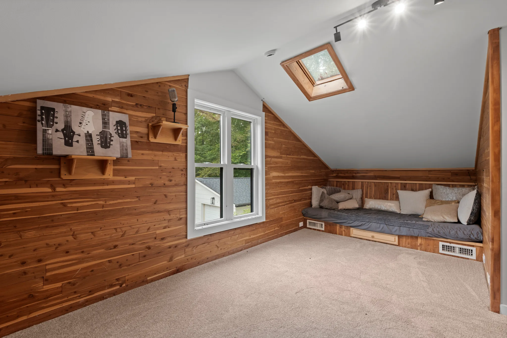Finished attic room with cedar plank walls, sloped white ceiling, skylight, carpeted floor and a built-in daybed with pillows