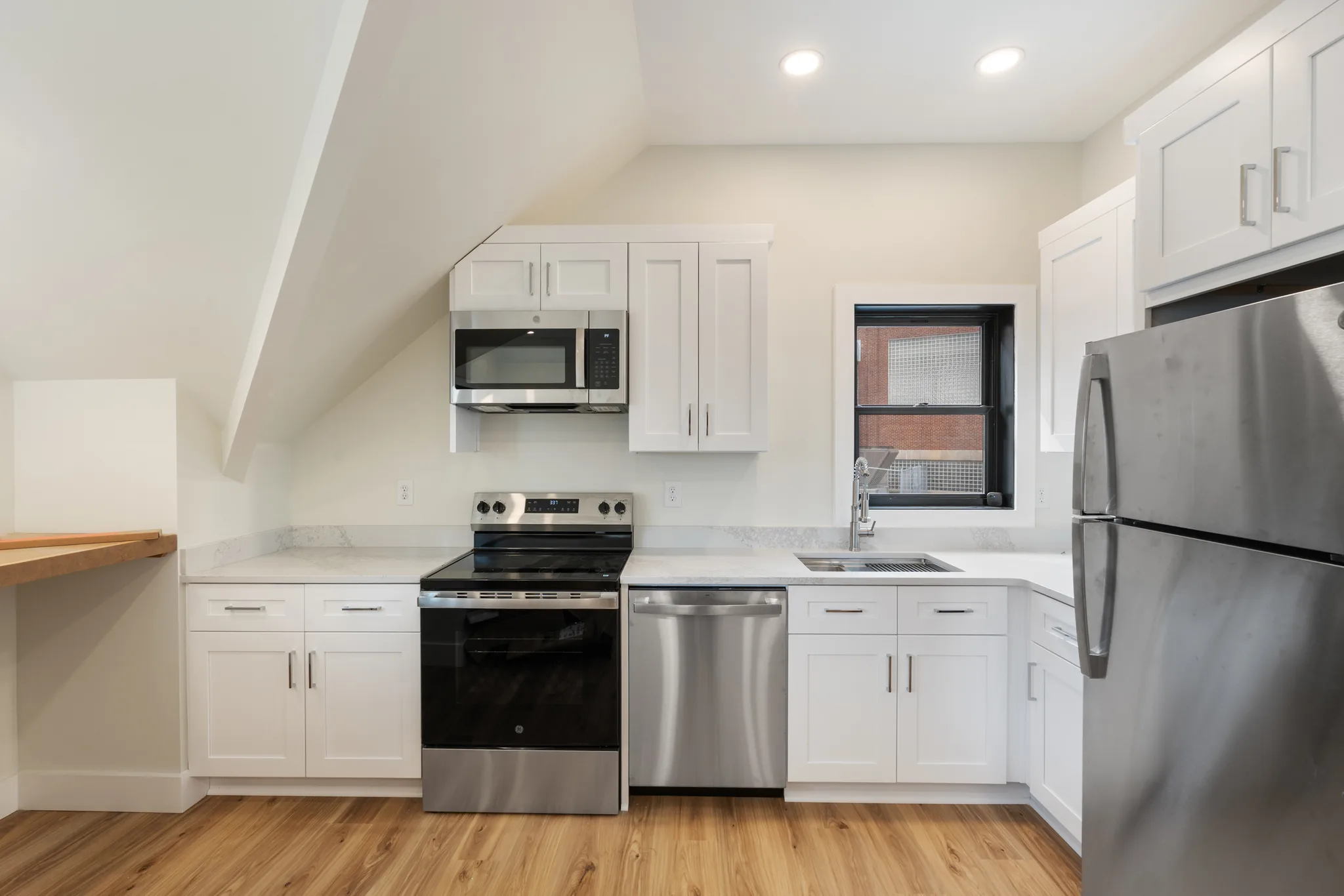 Compact kitchen with white shaker cabinets, light quartz counters, stainless range, dishwasher and refrigerator, sloped ceiling and wood-look plank flooring