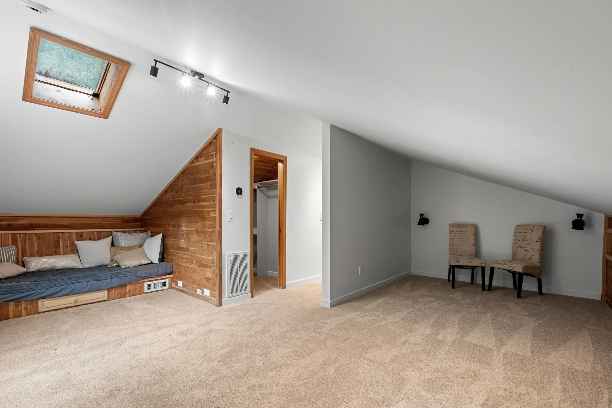 Finished attic room with sloped white ceilings, a wood-framed skylight, cedar-paneled built-in daybed, carpet, gray accent wall and two upholstered chairs