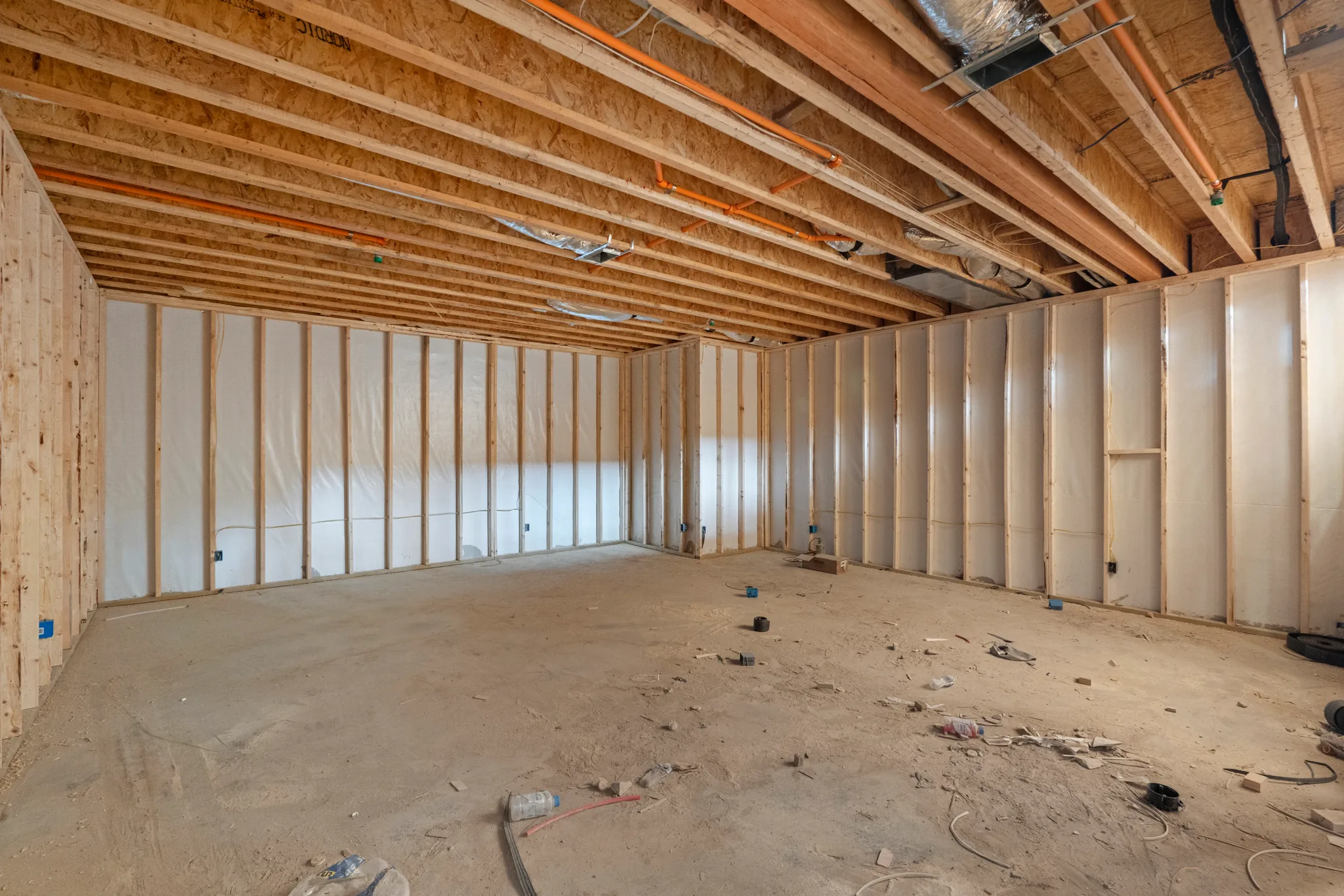 Unfinished basement room under construction with stud walls covered in white vapor barrier, exposed floor joists, ductwork and dirt floor