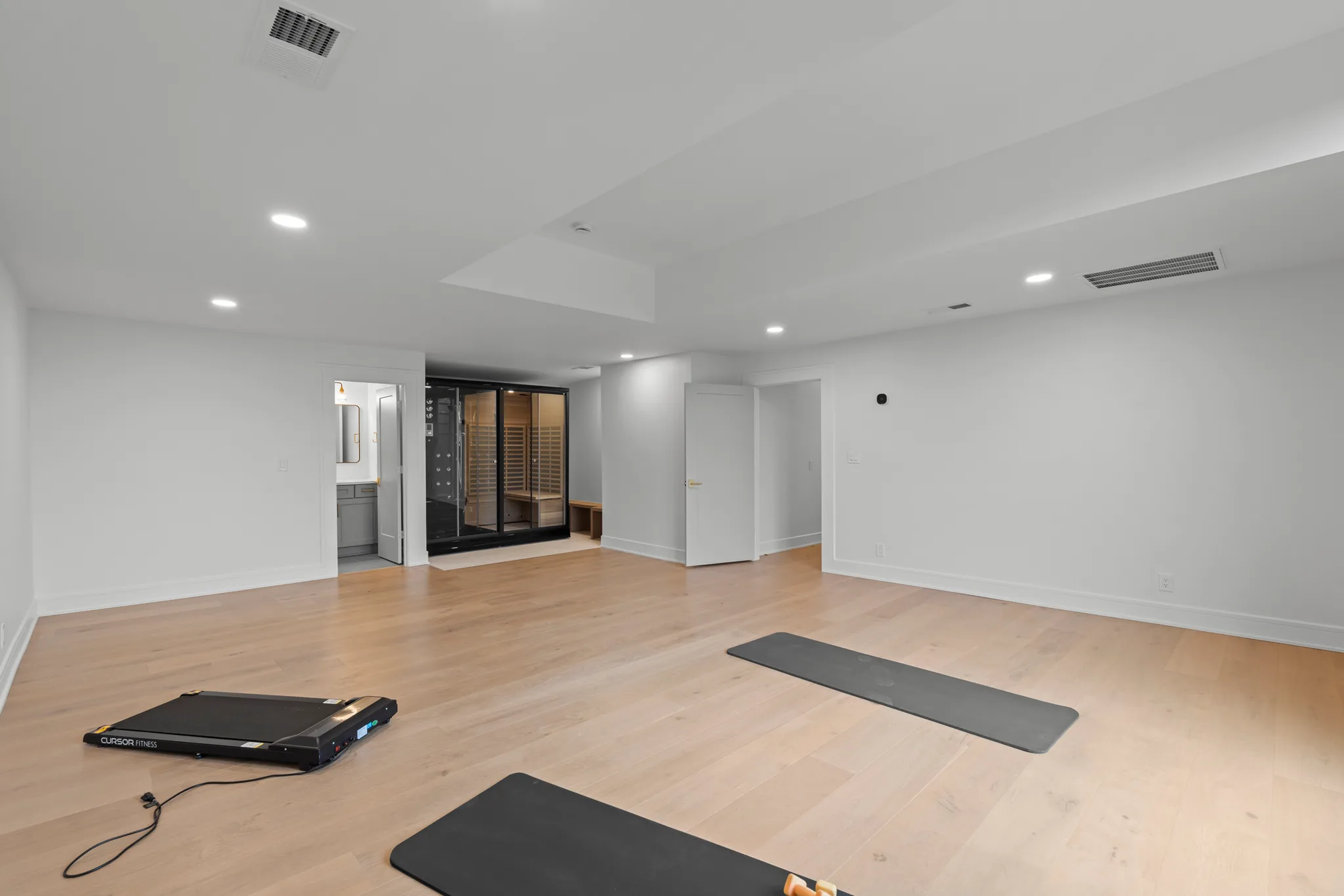 Finished lower level exercise room with light oak flooring, white walls, recessed lighting, exercise mats, folding treadmill, glass-front sauna and adjoining bathroom