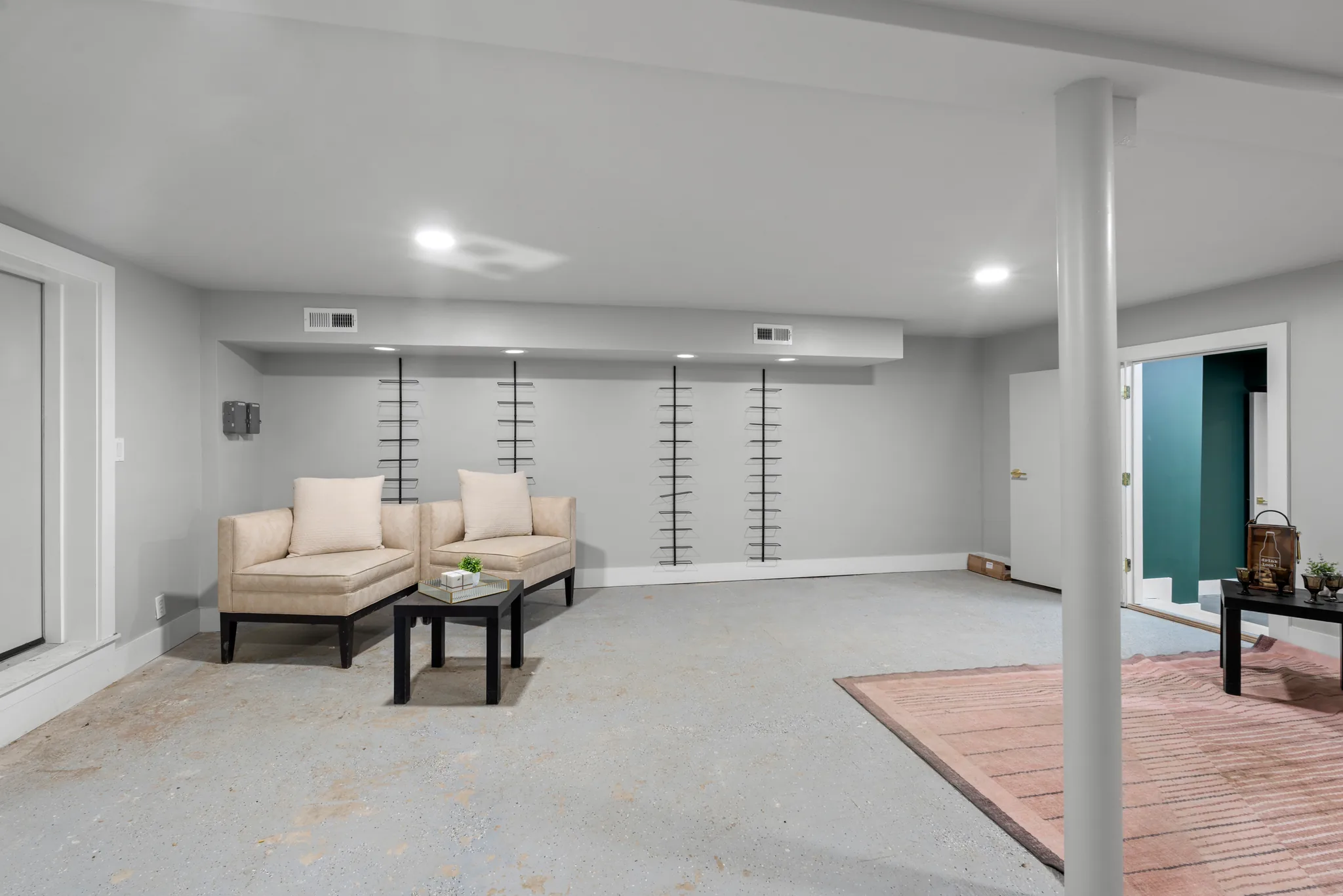 Basement remodel in progress with gray walls, recessed lighting, metal wall wine racks, two cream chairs and bare concrete floor