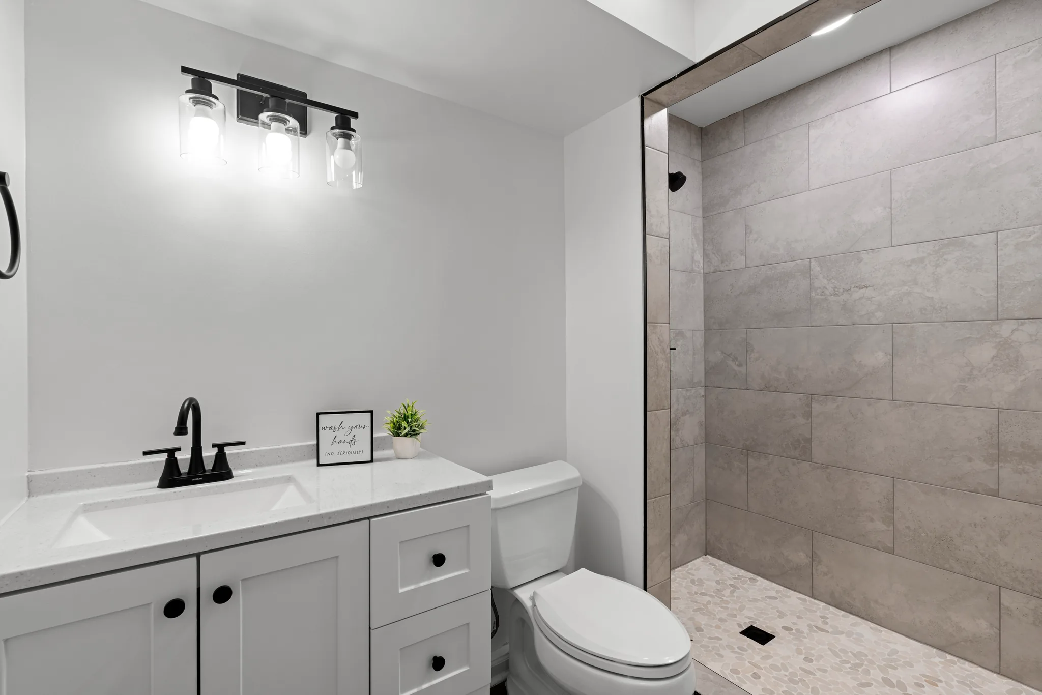 Bathroom with white shaker vanity, quartz top, black faucet and three-light fixture beside a walk-in shower with gray tile and pebble floor