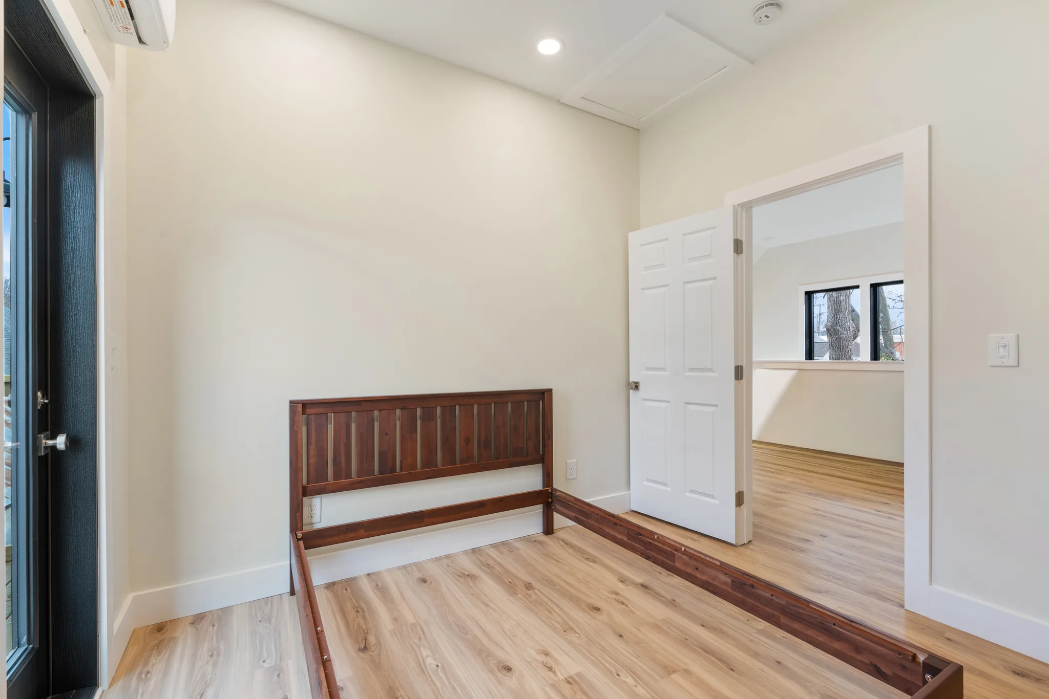 Empty bedroom with a dark wood bed frame, light plank flooring, white six-panel door open to an adjoining room, and a wall-mounted mini-split unit