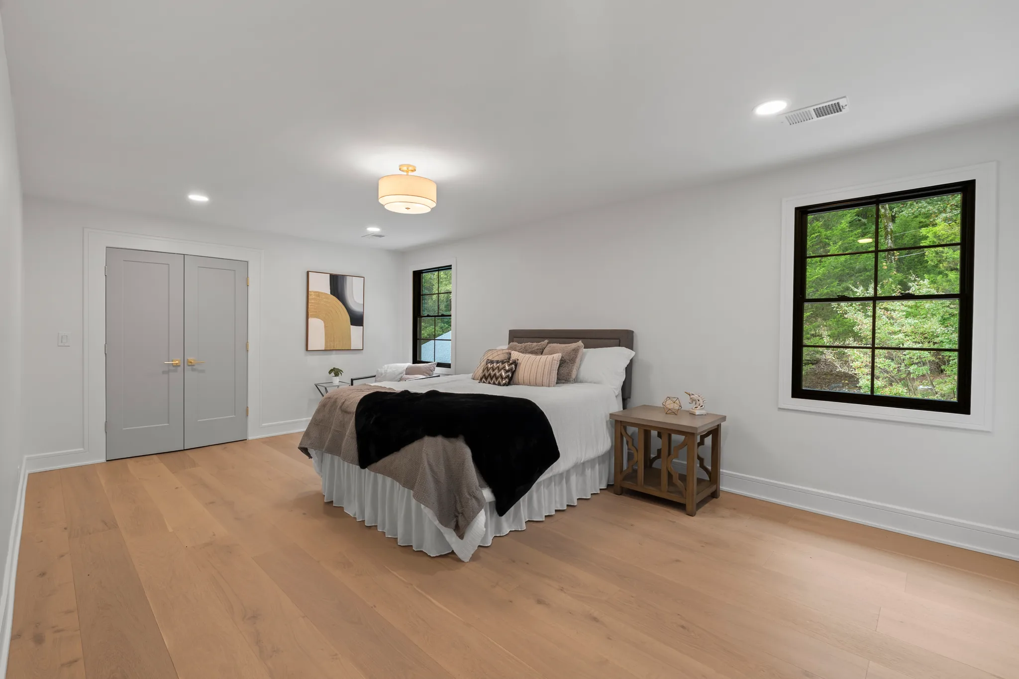 Bedroom with wide-plank oak flooring, white walls, gray double closet doors, black-framed window looking into green woods, and a drum ceiling light