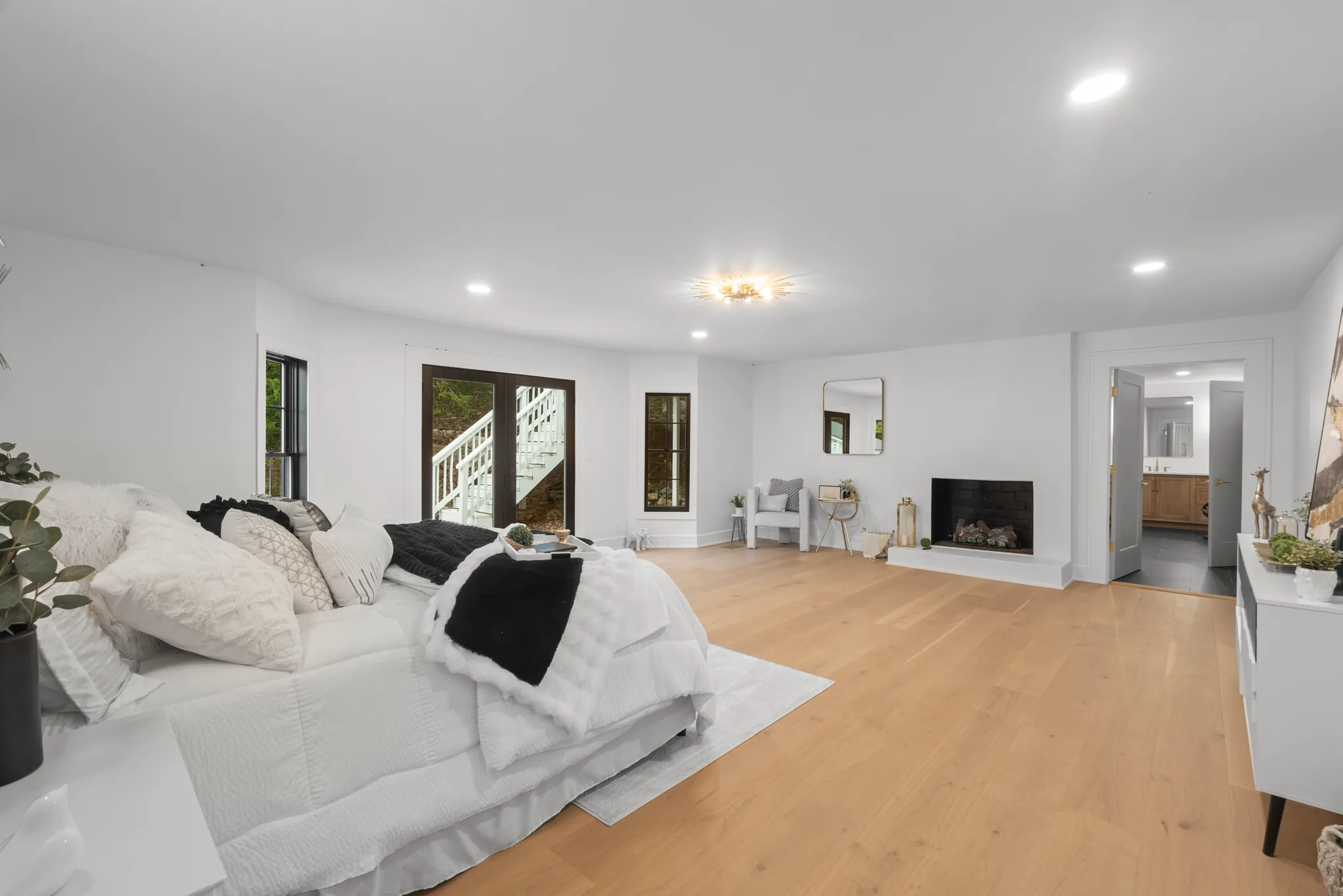 Spacious white bedroom with light oak plank floors, a black-surround corner fireplace, French doors to an exterior stair, and a doorway to a bath